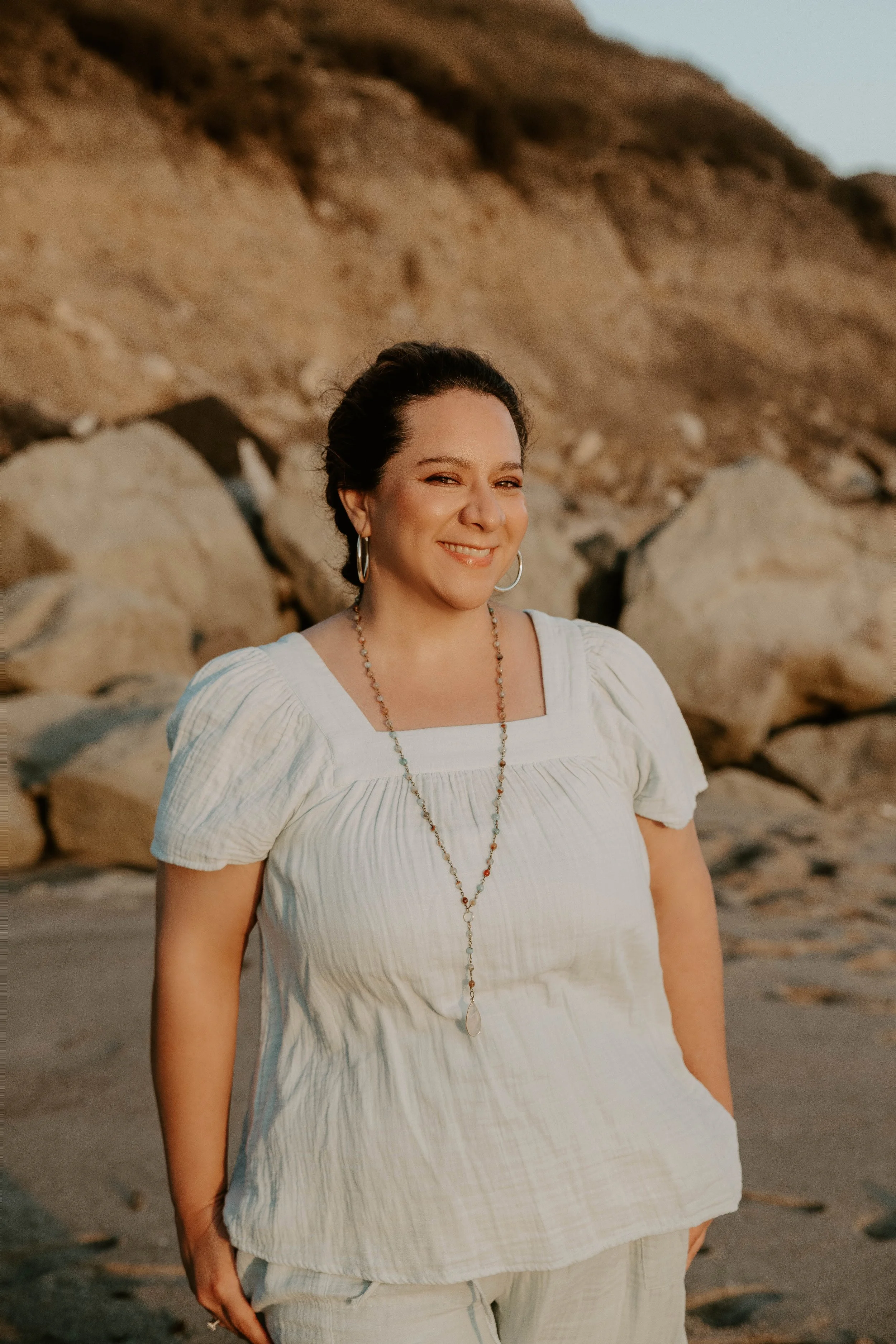 A woman with dark hair braided back, wearing a white blouse and beige pants, standing on a rocky beach with large rocks and a hill in the background, smiling at the camera.