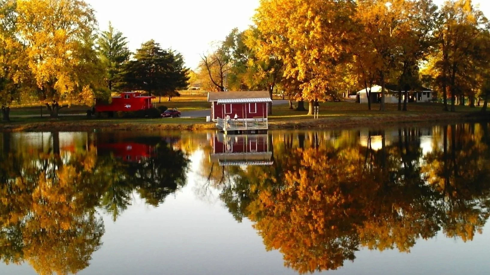 Autumn scene showing a pond with reflections of trees with yellow and orange leaves, a small red boathouse, a red shed, and surrounding houses.