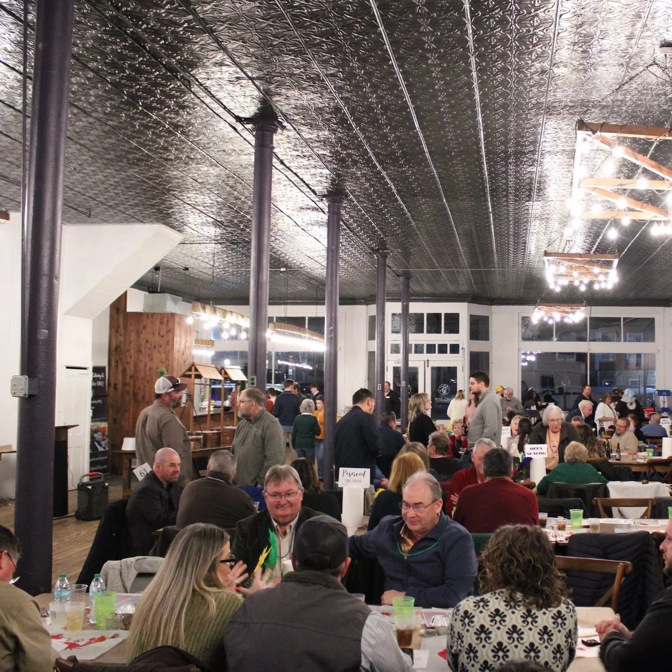 A large gathering of people dining and socializing in a spacious, well-lit restaurant with high ceilings, black metal support columns, and wooden accents.