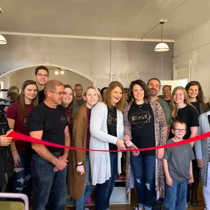 Group of people gathered indoors for a ribbon-cutting ceremony, with woman in white cardigan cutting a red ribbon.