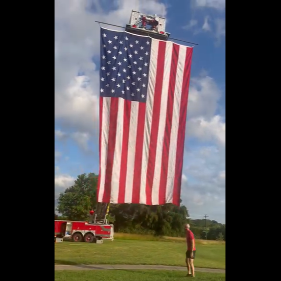 A large American flag hanging vertically from a fire truck ladder extended in the air during daytime, with a person standing on the ground watching.