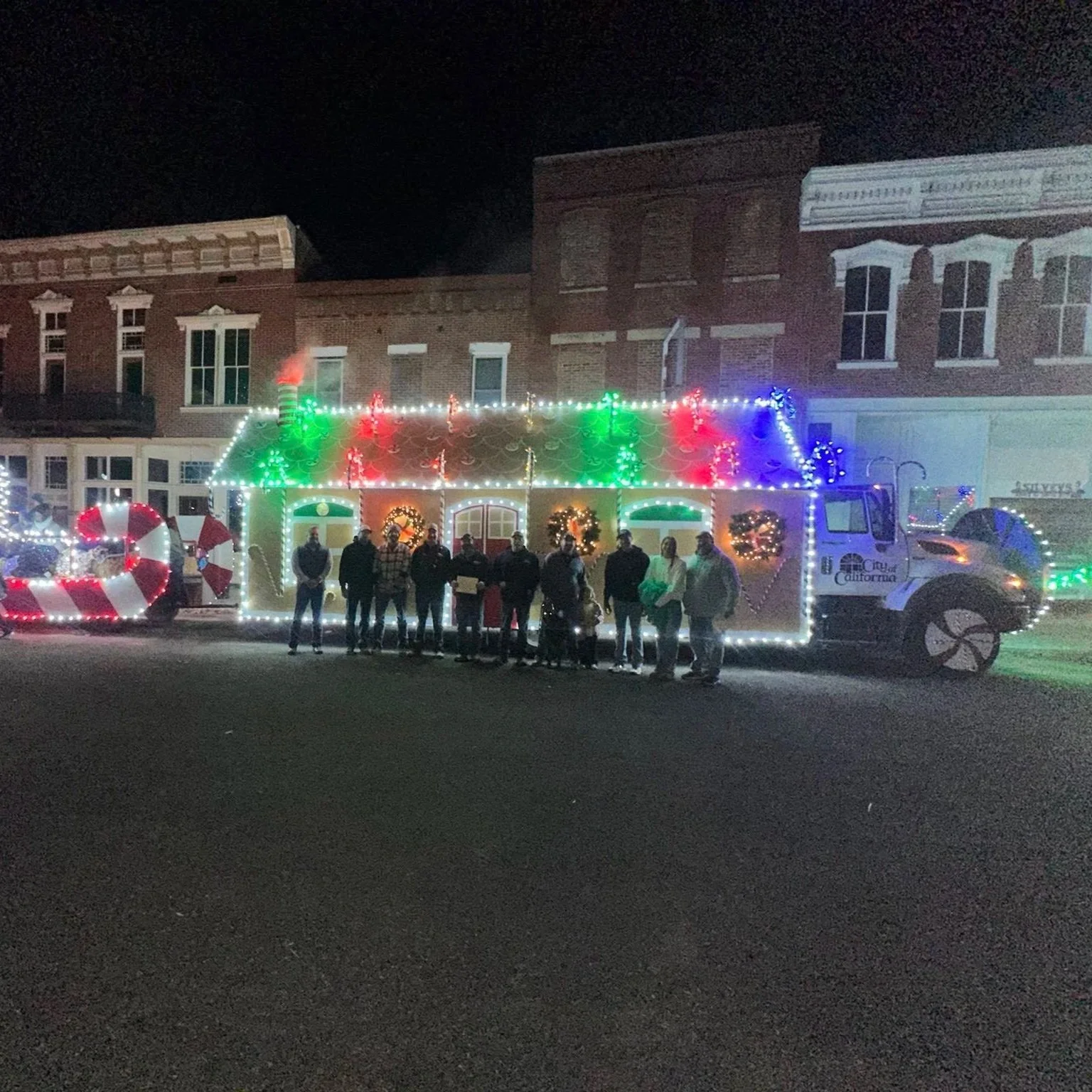 A decorated Christmas float shaped like a house with Christmas lights, wreaths, and candy cane decorations, and a group of people standing in front of it during nighttime.