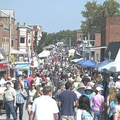 Crowd of people walking down a busy Main Street lined with tents and shops during an outdoor event.