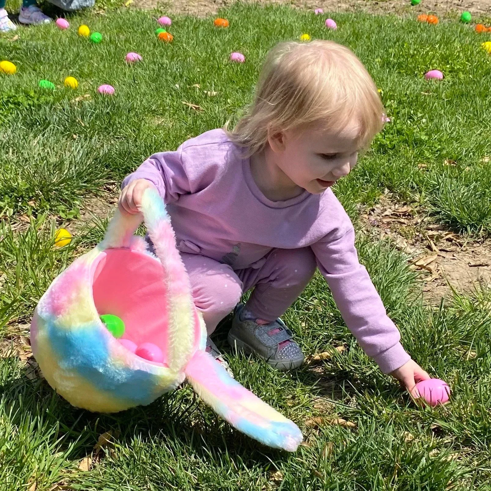 A young girl with blonde hair, wearing a lavender shirt and lavender pants, playing outdoors on grass with Easter eggs, holding a colorful, fluffy Easter basket.