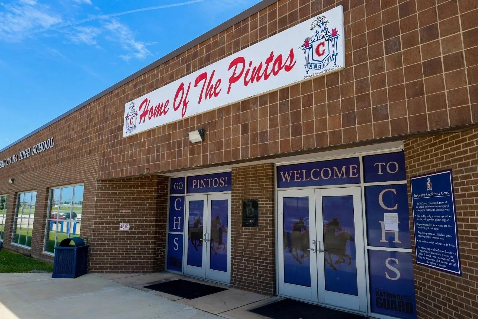 Front entrance of a high school building with a brick exterior, large signs reading 'Home of The Pintos' and 'Welcome to CHS', and glass doors with pinto horse images. There is a trash can outside, windows reflecting the parking lot and sky, and informational signs near the doors.