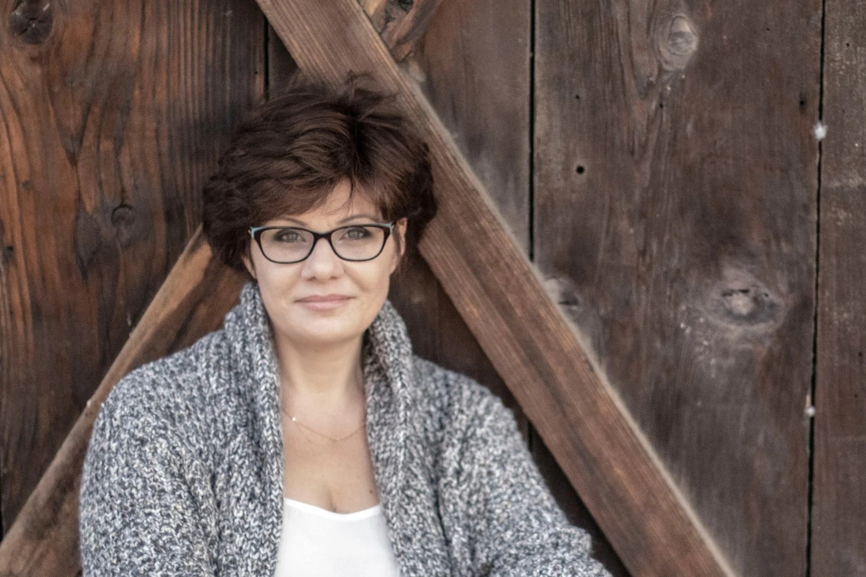 A woman  writer with short brown hair, wearing glasses, a gray sweater, and a white top, sitting and smiling in front of a wooden wall.