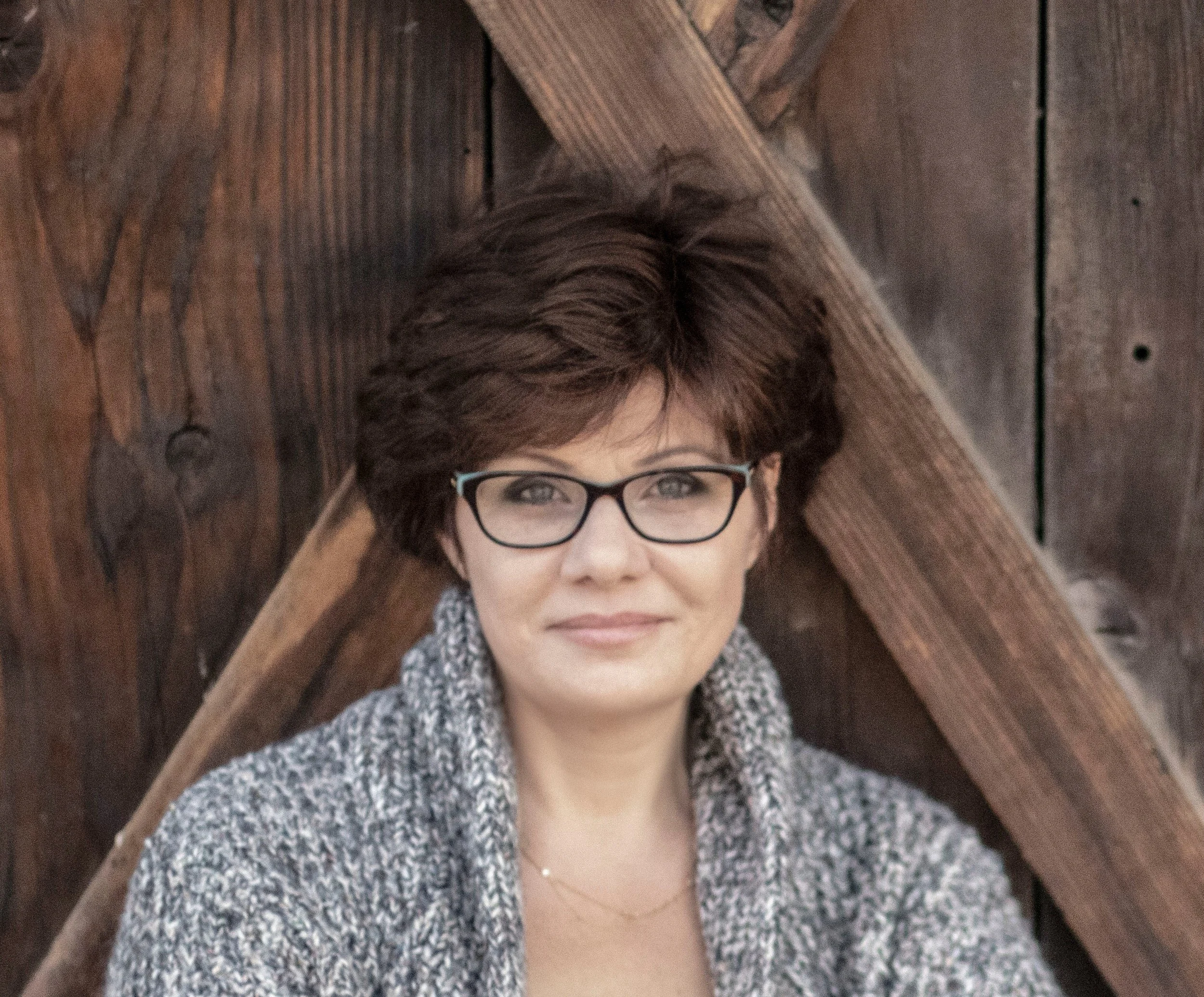 A woman with short brown hair and glasses lies on a wooden floor, looking up at the camera.
