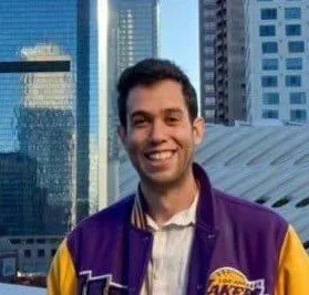 A young man smiling in front of skyscrapers outside on a city street, wearing a purple and yellow jacket.