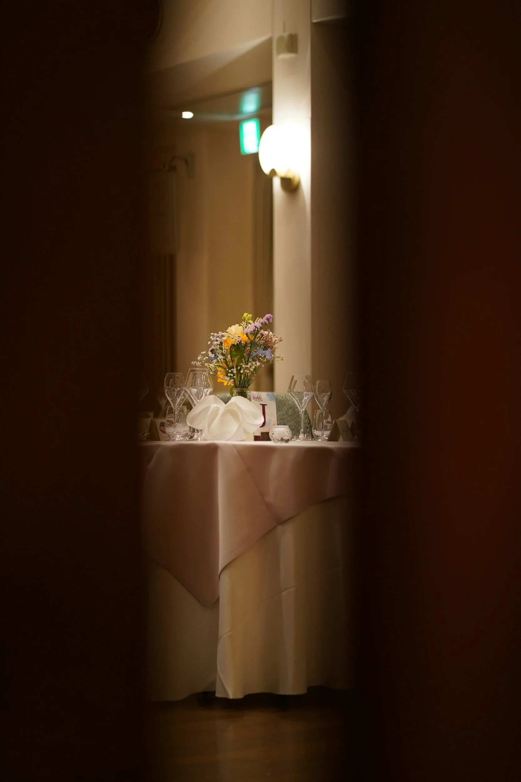Een gedekte tafel met een bloemstuk, wijnglazen en een tafelnummerkaart, gezien door een kleine opening of opening in een muur in een restaurant of banquetruimte.