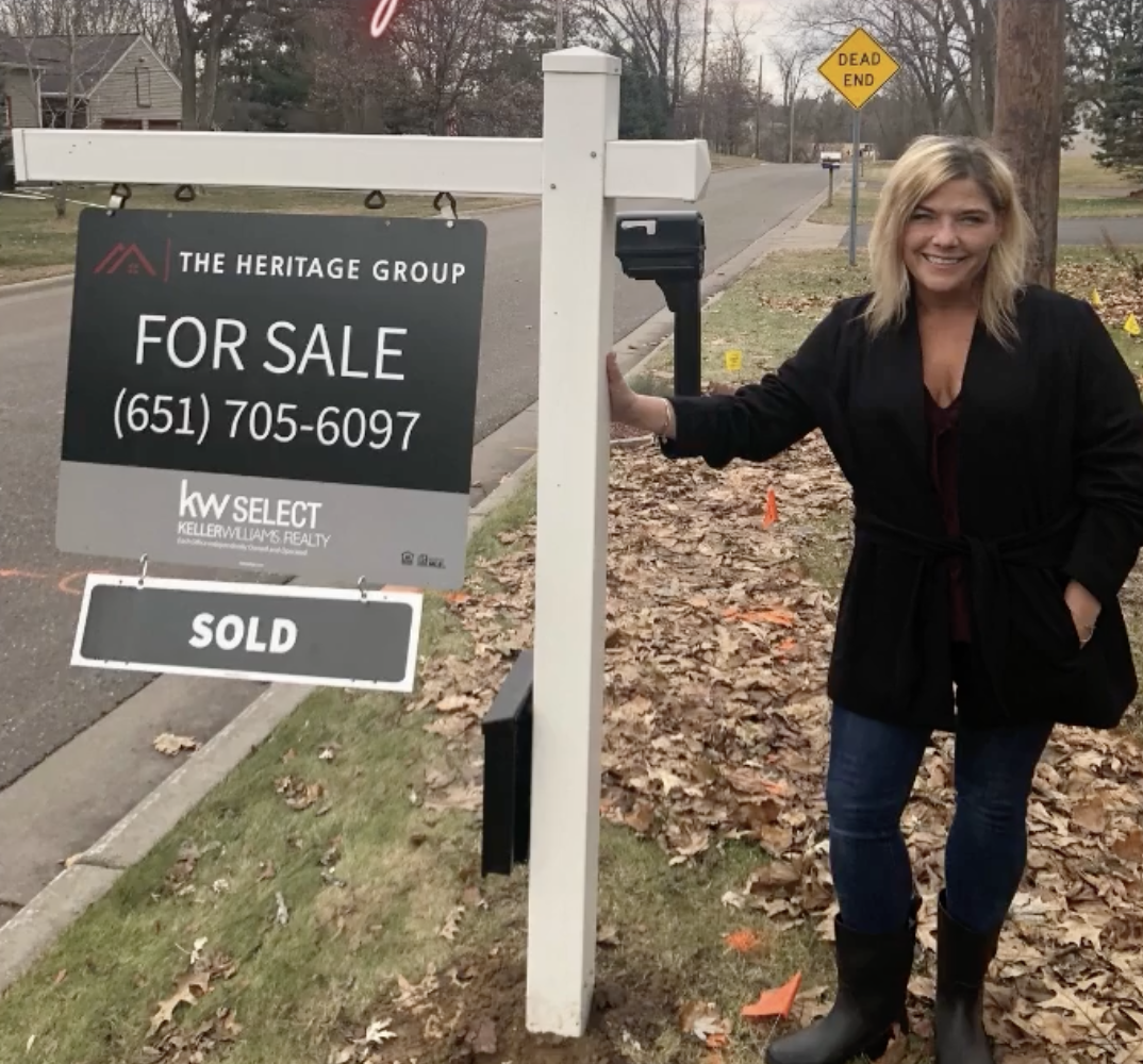 A woman standing beside a 'For Sale' sign on a suburban street, smiling at the camera. The sign indicates the property has been sold.