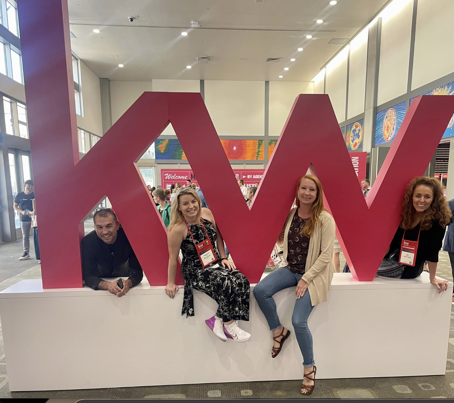 Four people leaning on a large red letters spelling 'KW' at a conference or event in a spacious, well-lit indoor venue with other attendees in the background.