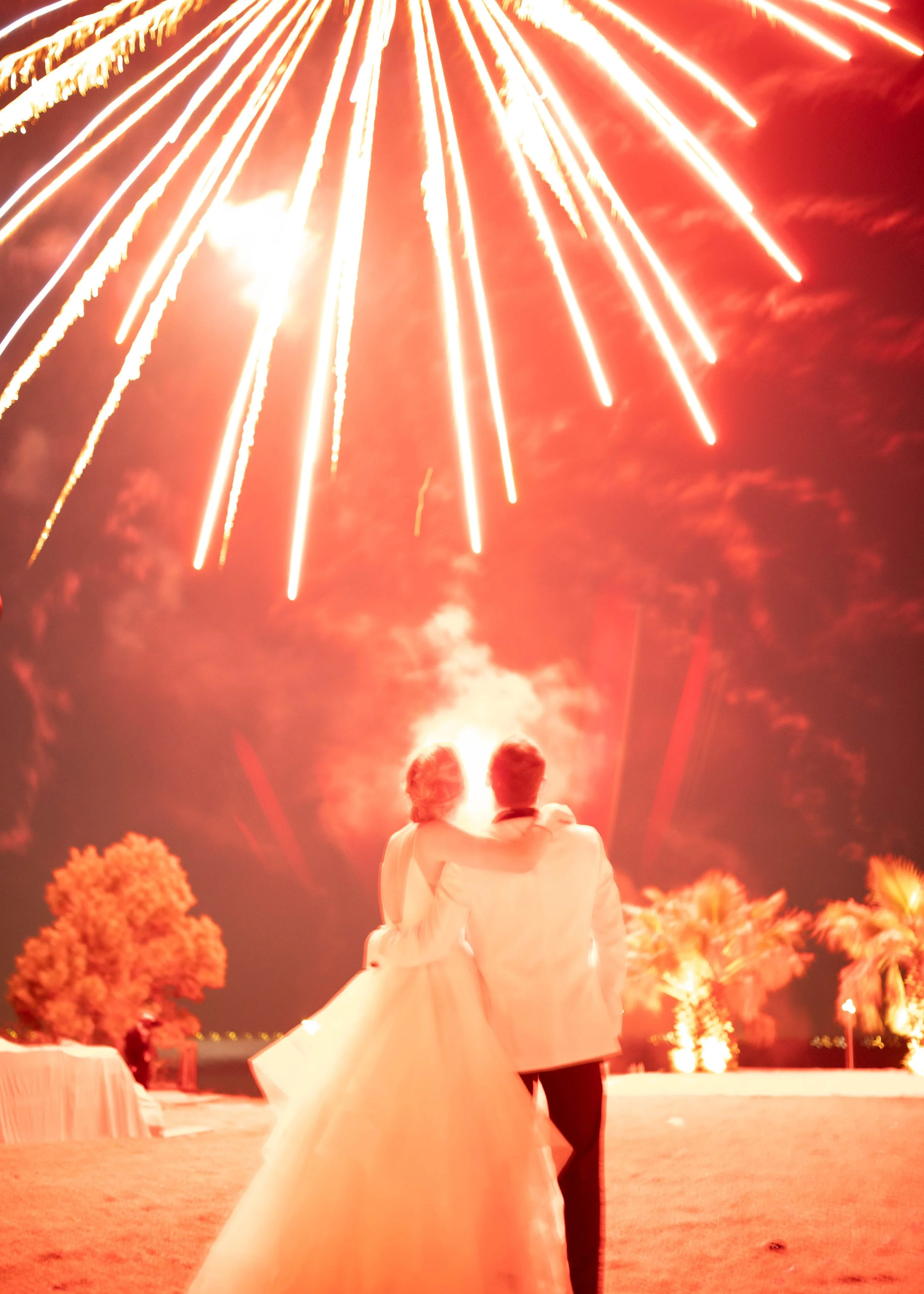 A newly married couple in white wedding attire watching a fireworks display at night.