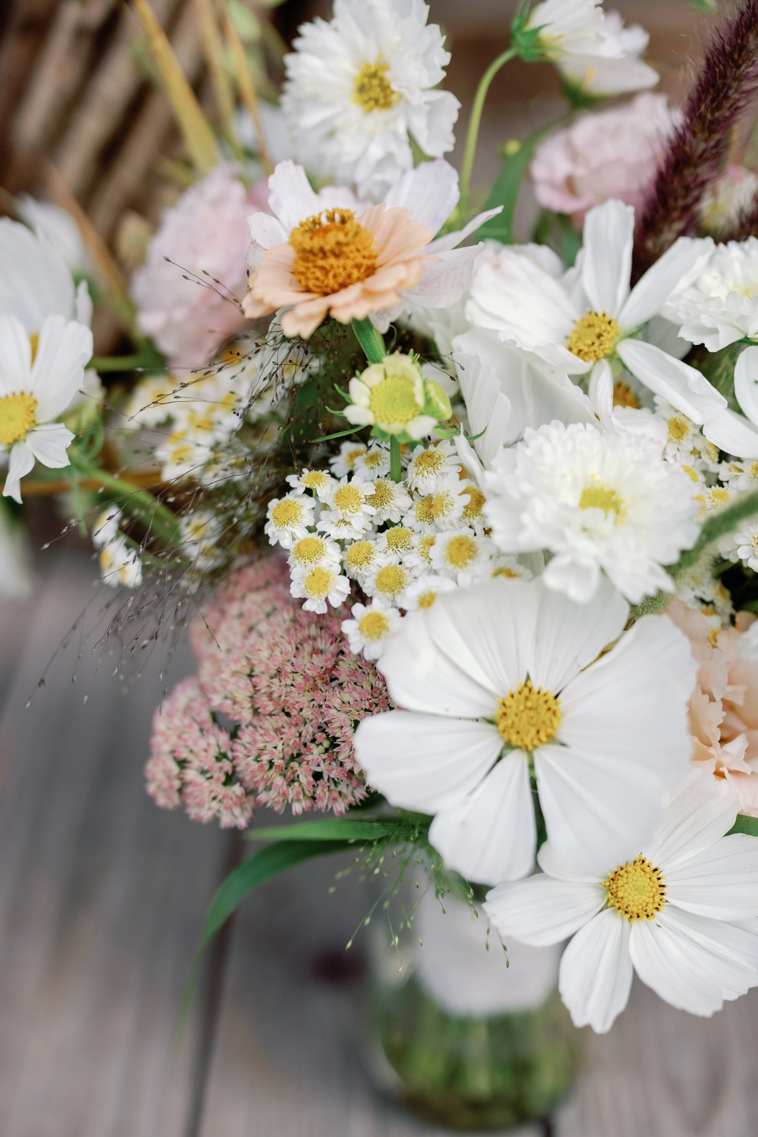 A close-up of a colorful mixed flower bouquet in a glass vase, featuring white, pink, and yellow flowers with greenery on a wooden surface.