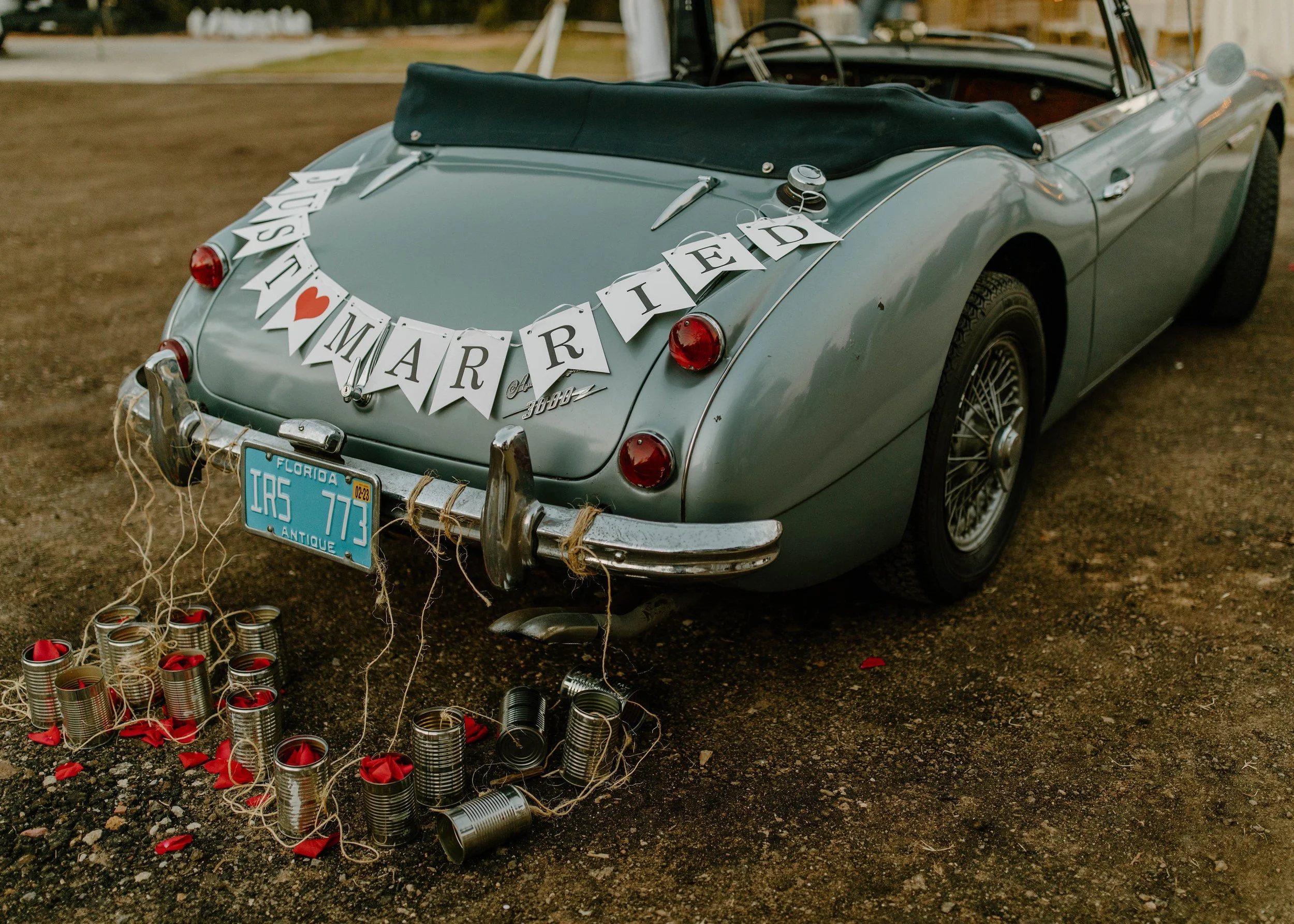 Vintage light blue convertible car decorated for a wedding with a banner that reads 'Just Married' and a small red heart. The car has a Florida license plate, and cans tied with string are attached to the rear bumper, with some cans and small red ite