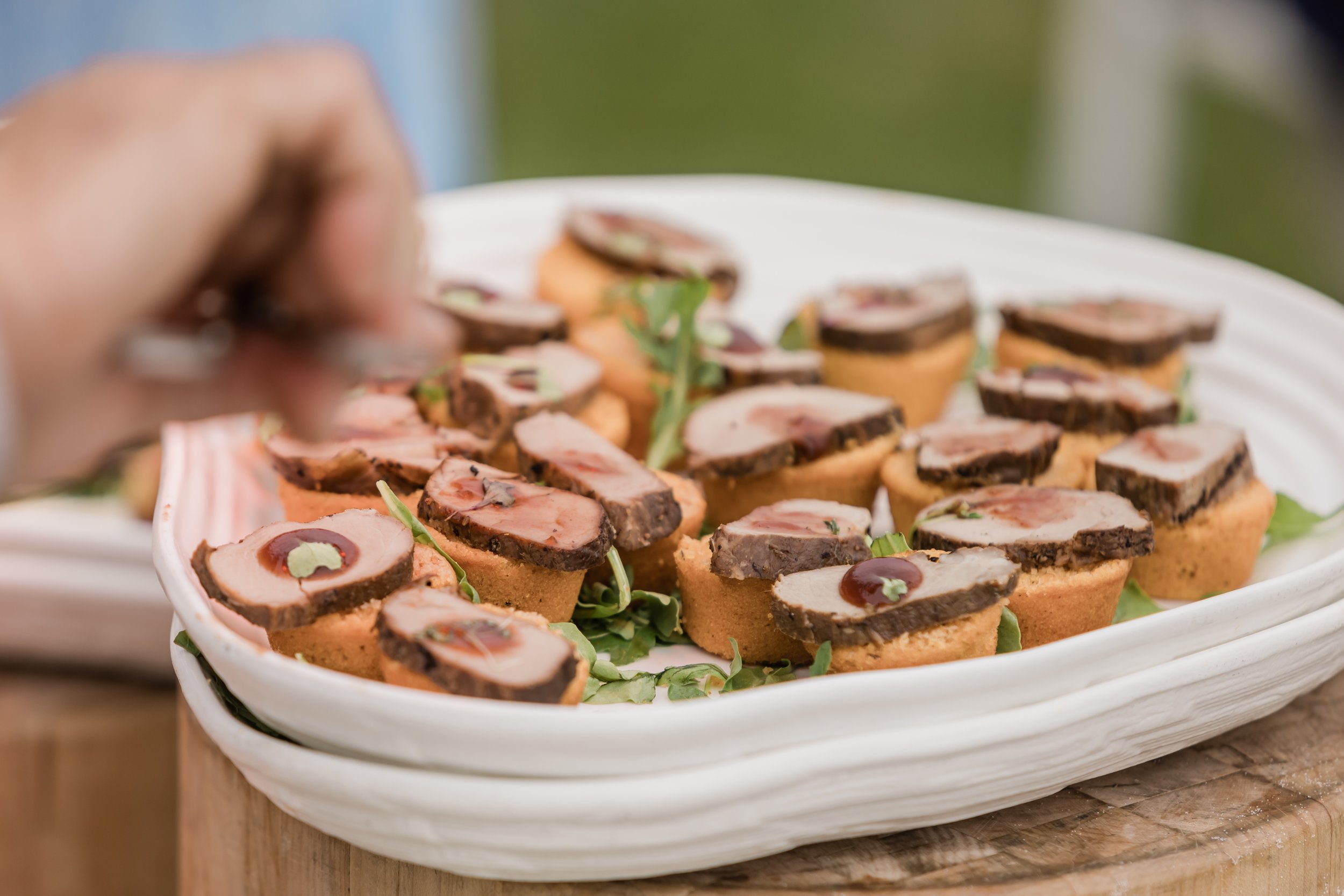 Close-up of a white platter filled with small appetizers made of sliced meat on a crispy base, garnished with a small amount of sauce and microgreens, on a wooden surface outdoors.