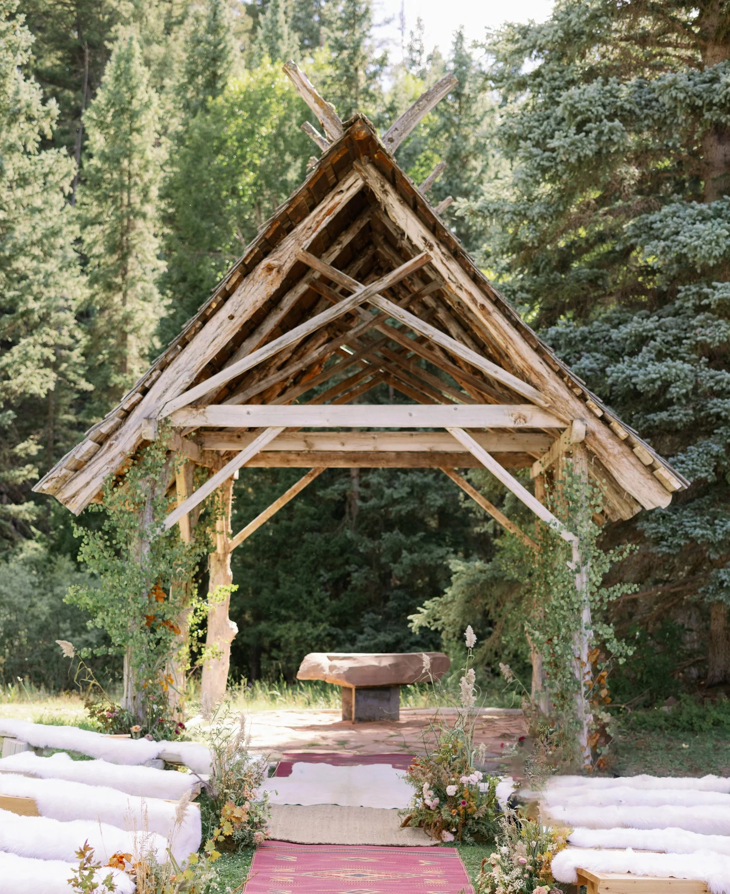 A rustic outdoor wedding ceremony setup with a wooden arch made of raw, weathered wood, decorated with greenery and flowers, set in a forest clearing with tall trees in the background.