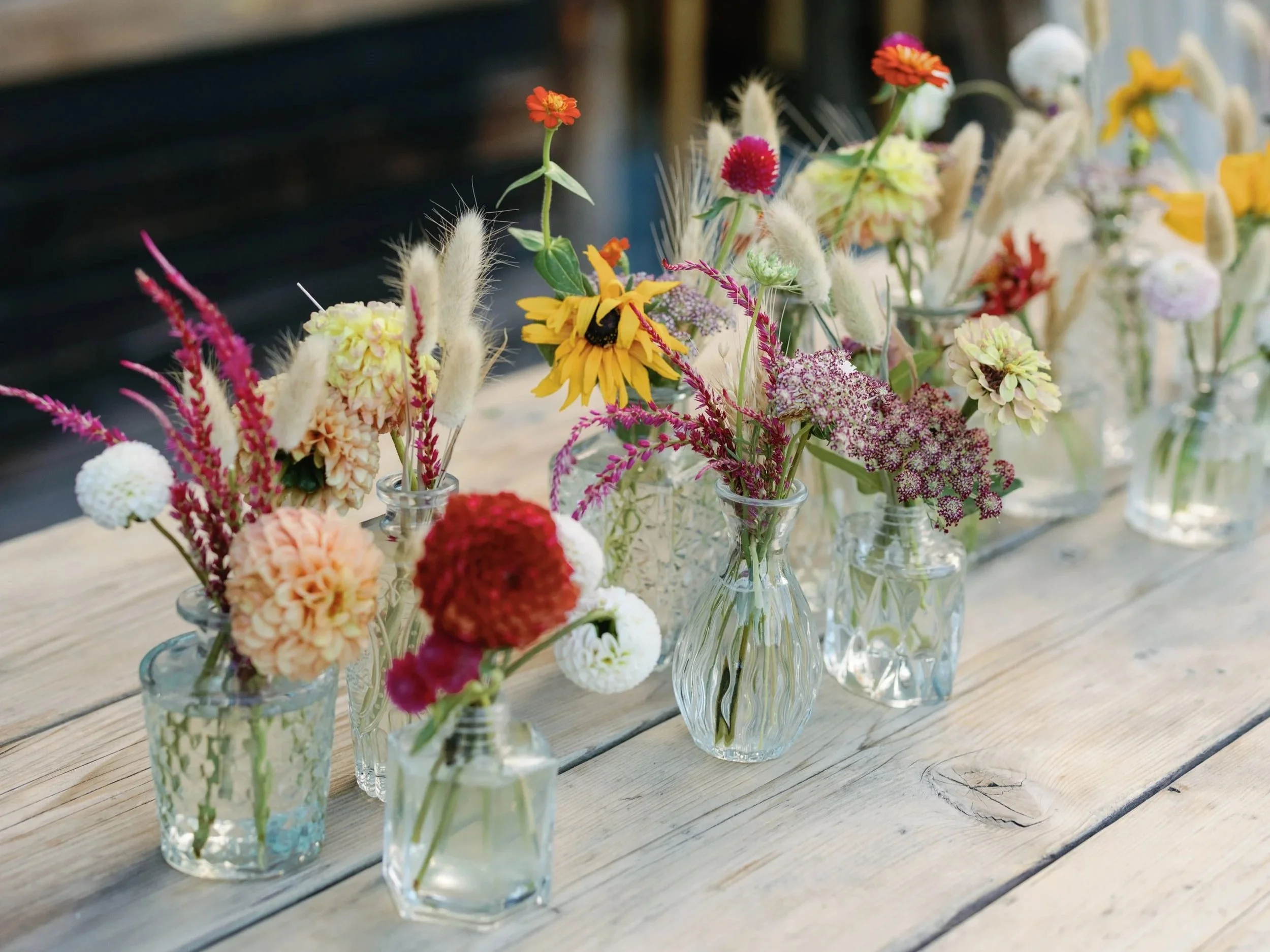 Various colorful flowers in clear glass vases arranged on a wooden table.