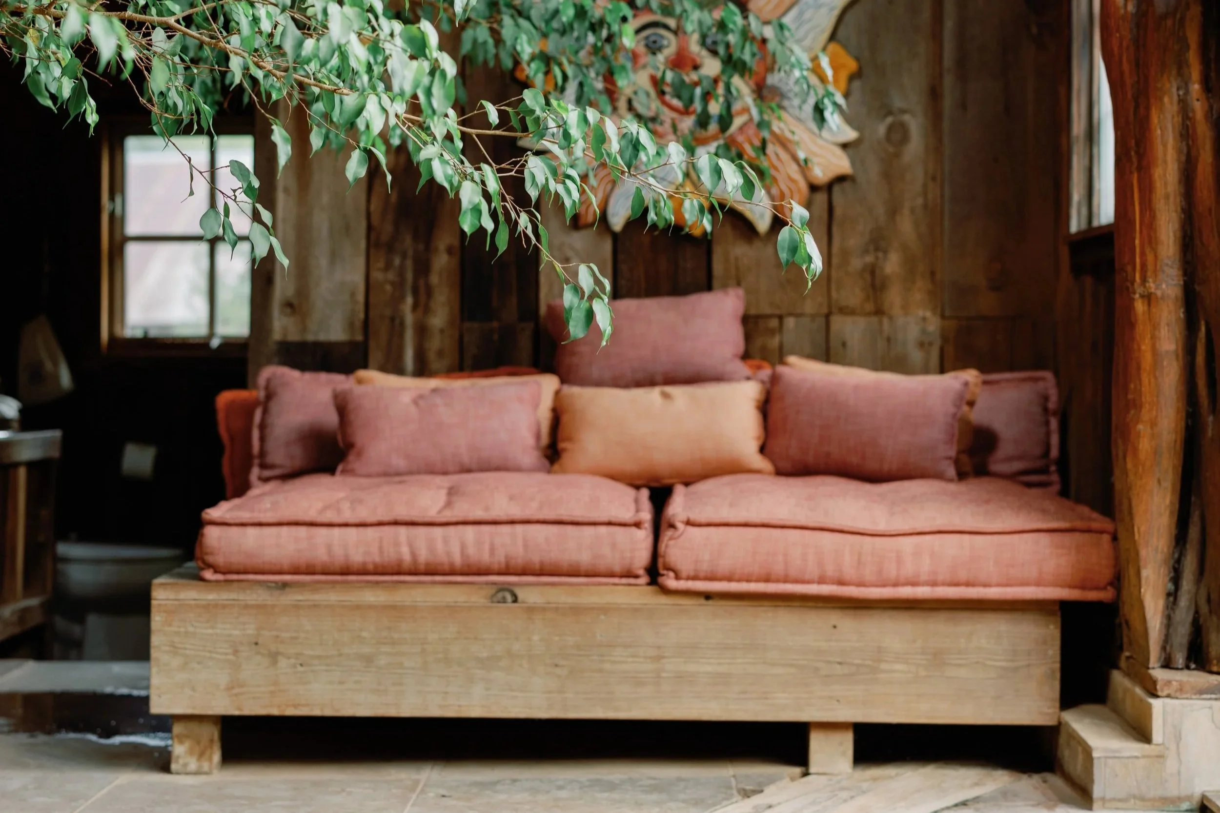 A rustic wooden room features a pink upholstered sofa with multiple cushions, a green leafy plant hanging from the ceiling, walls decorated with colorful tribal masks, and a window in the background.