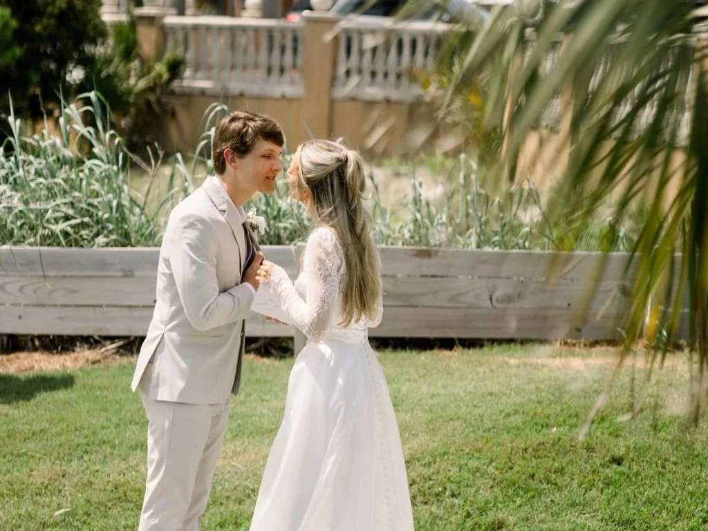 A couple, dressed in wedding attire, stand close together outdoors on a grassy area, gazing into each other's eyes with smiles, surrounded by greenery and trees.