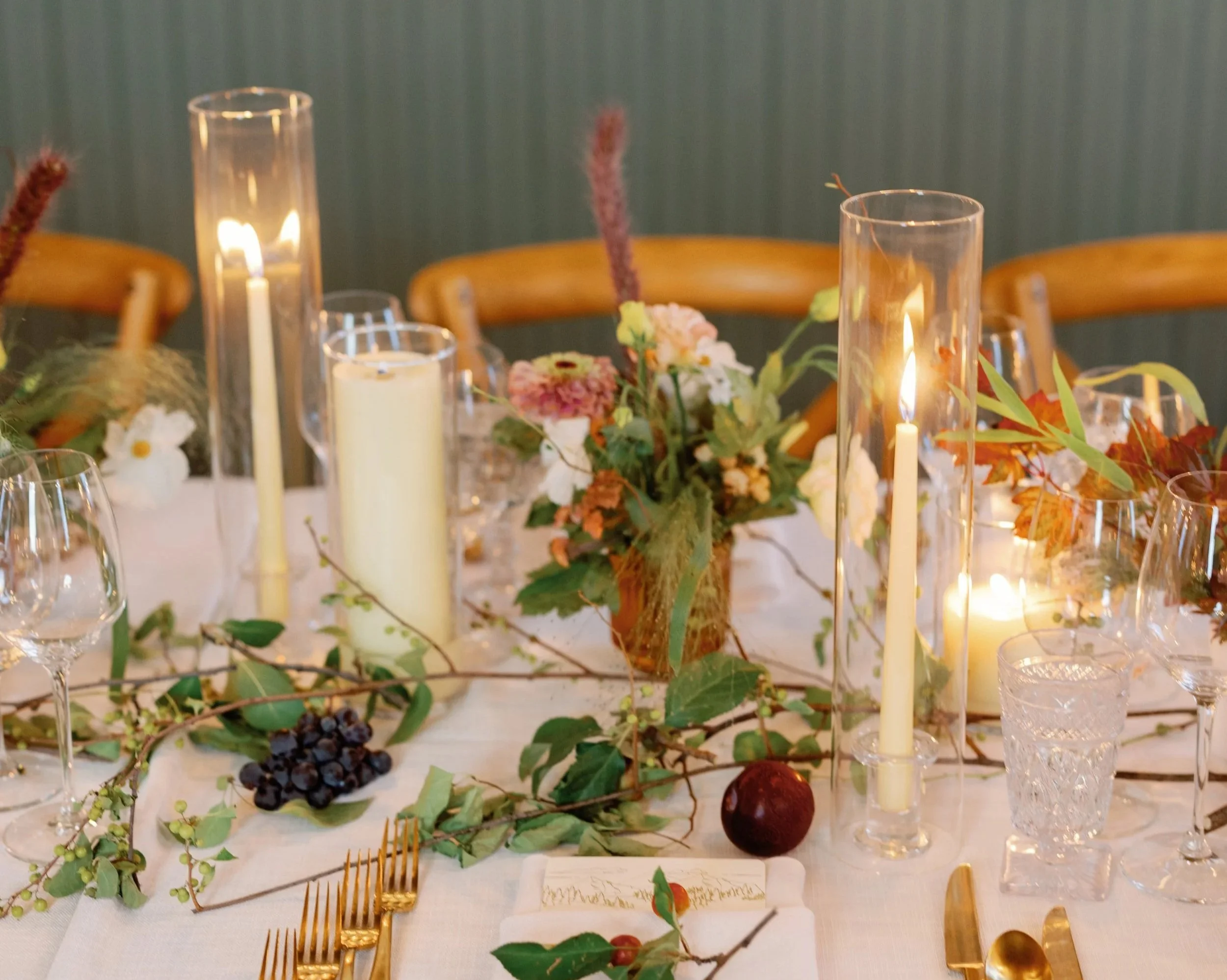 A decorated dining table with candles, floral arrangements, grapes, and greenery.