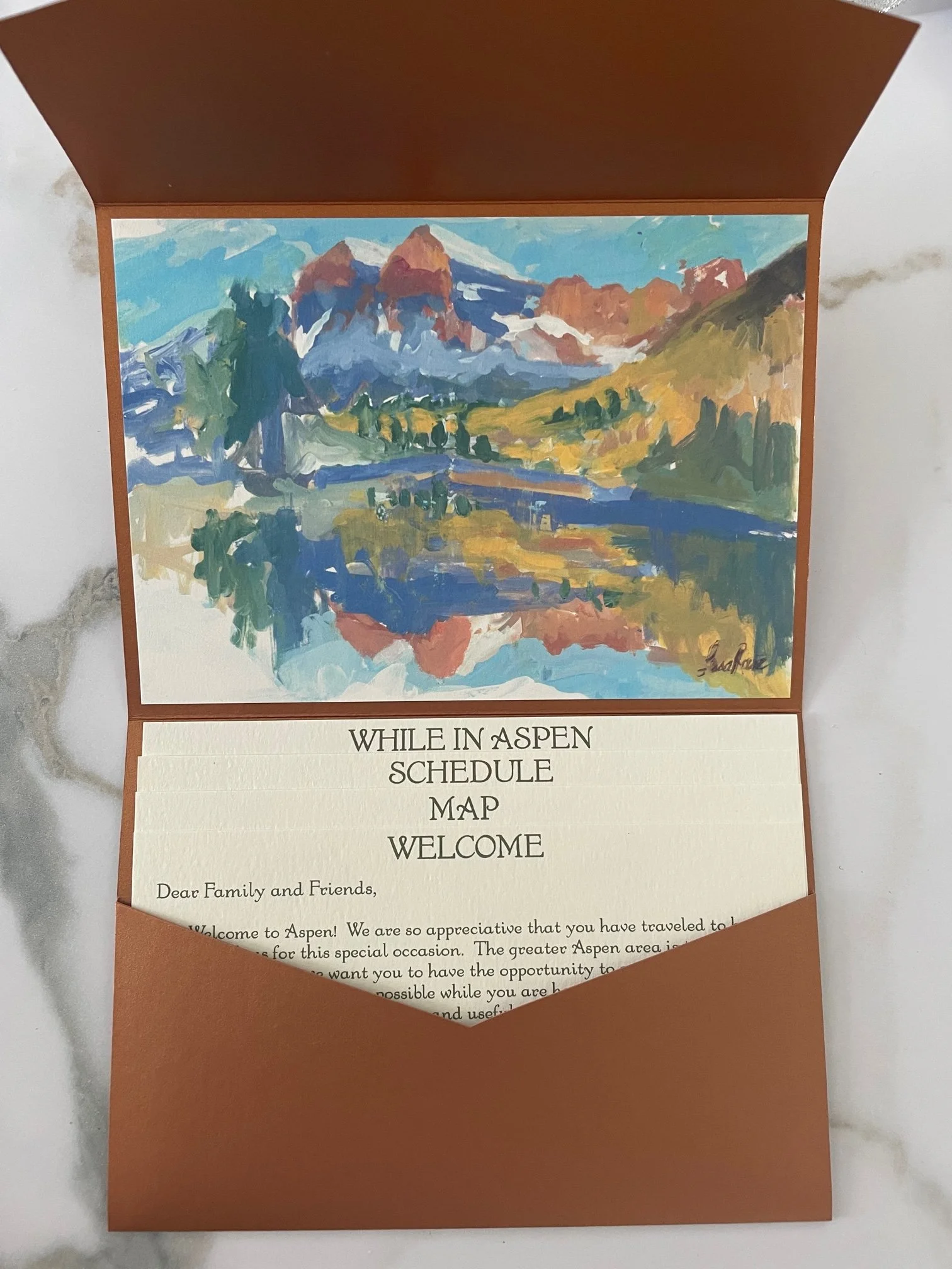 A printed invitation with an artistic painting of mountains, a river, and trees in shades of blue, green, yellow, and brown, placed inside a brown envelope.