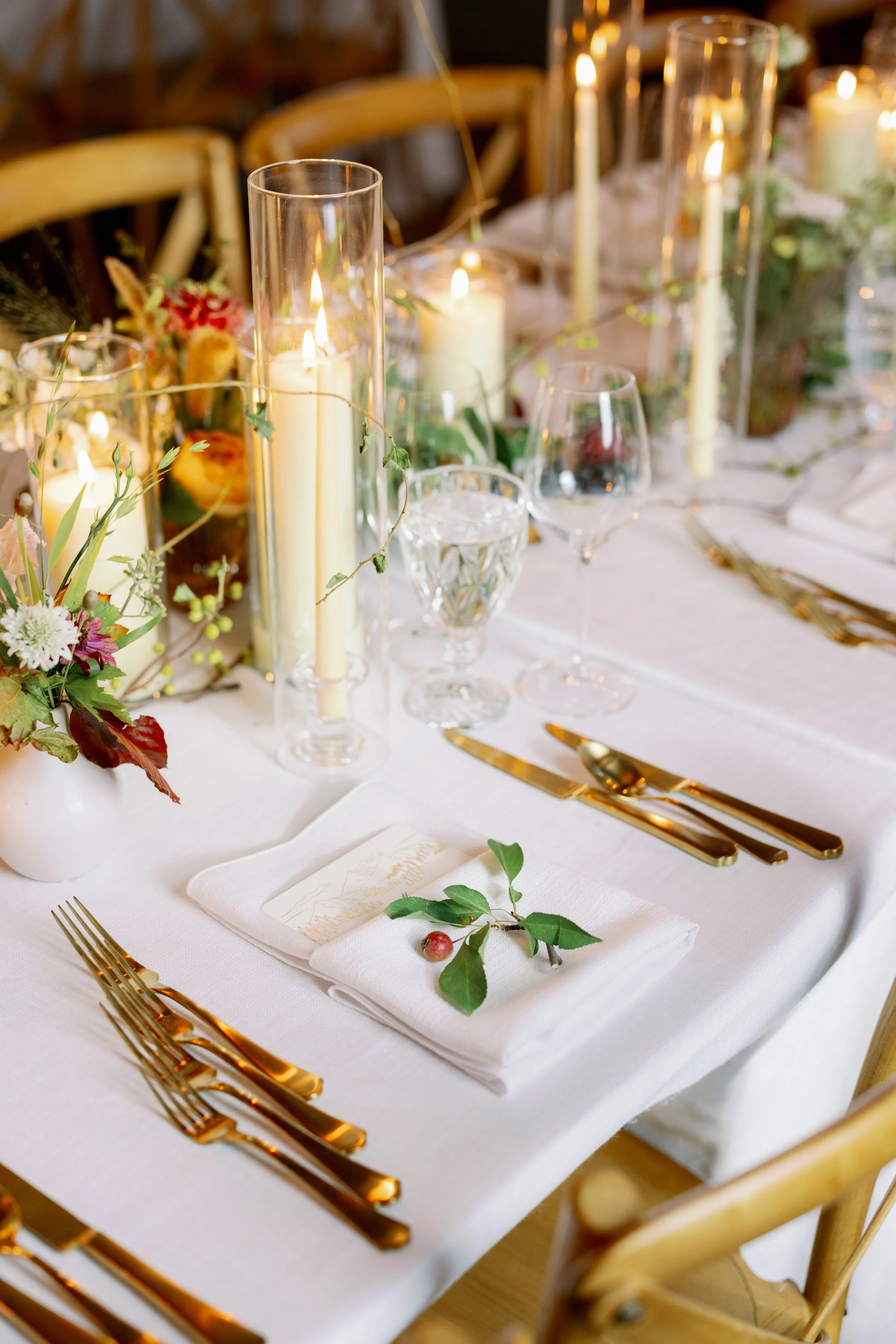 Elegant dining table set with white tablecloth, gold cutlery, glassware, and candles, decorated with flowers and greenery.