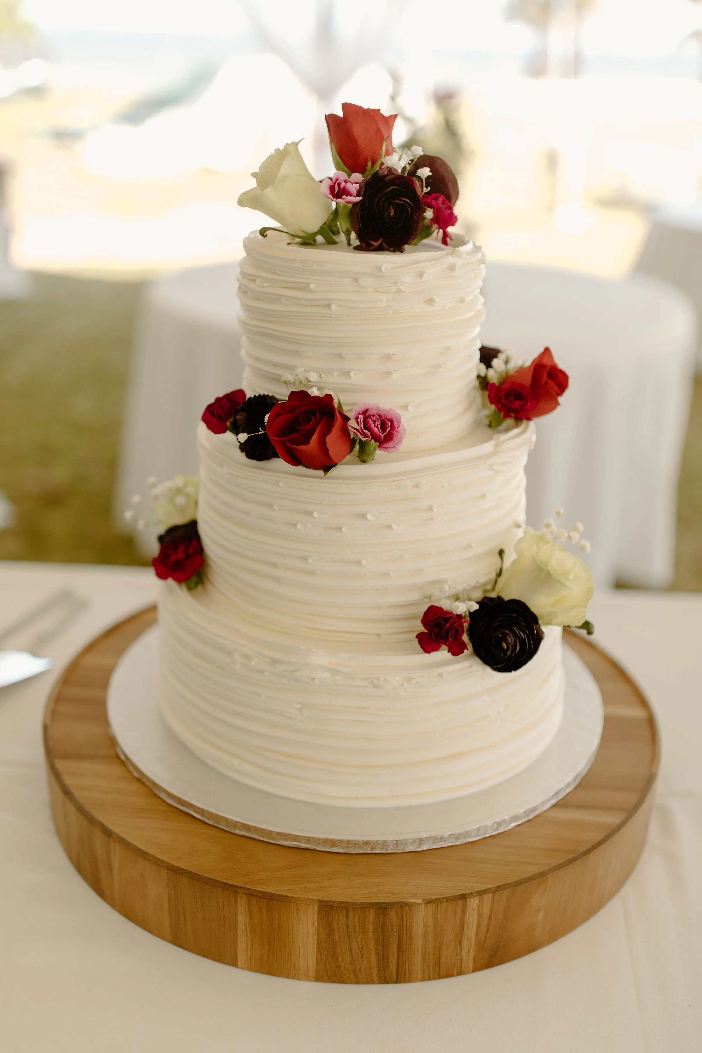 Three-tier white wedding cake decorated with fresh red, pink, black, and white flowers on a round wooden cake stand.