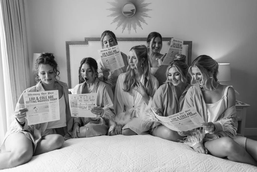 Six women in robes sitting on a bed, reading newspapers about a wedding, with one woman in the center and four women behind her, in a bedroom with a mirror and lamps.