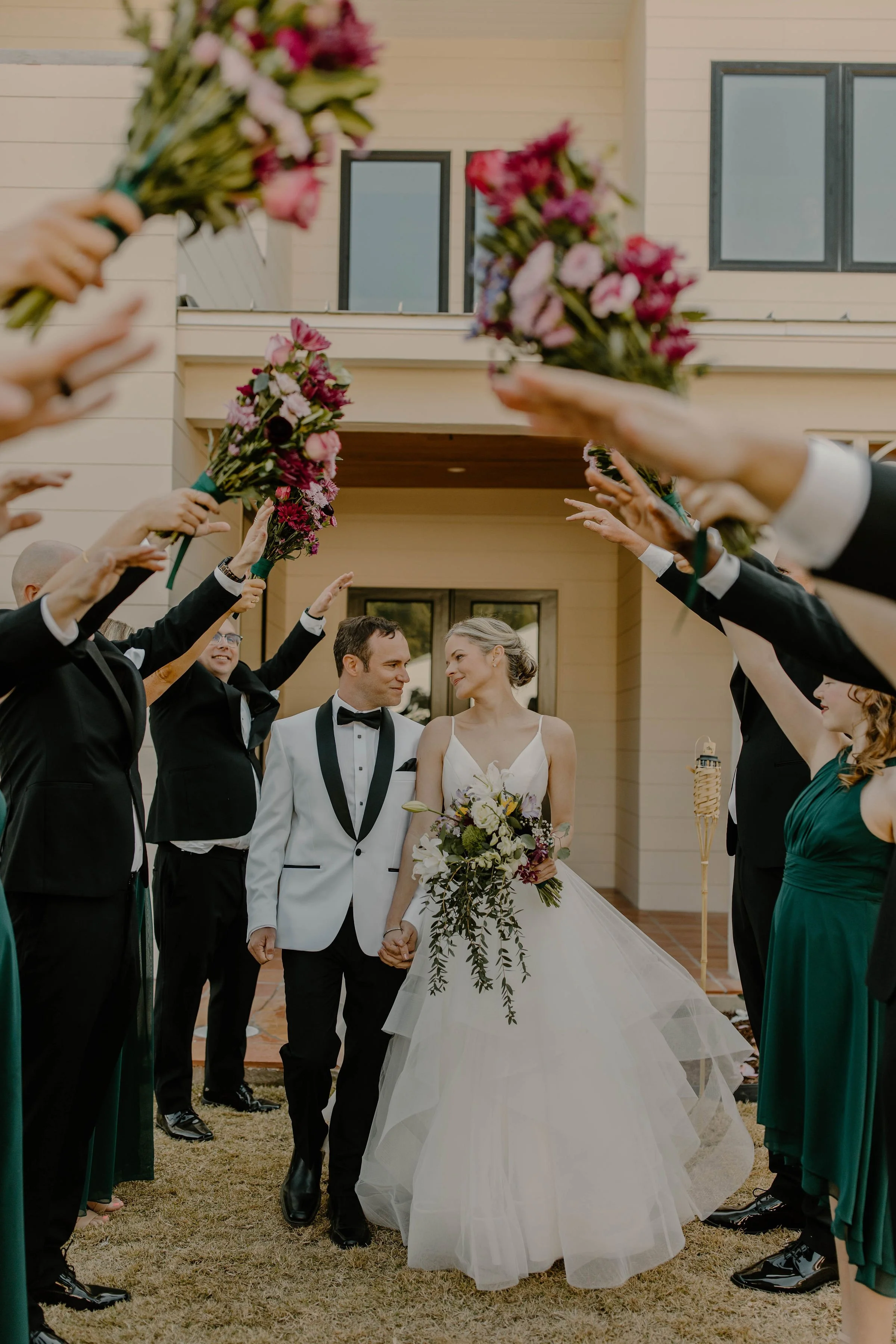 Bride and groom holding hands, walking through a tunnel of wedding guests holding bouquets, outside a modern house.