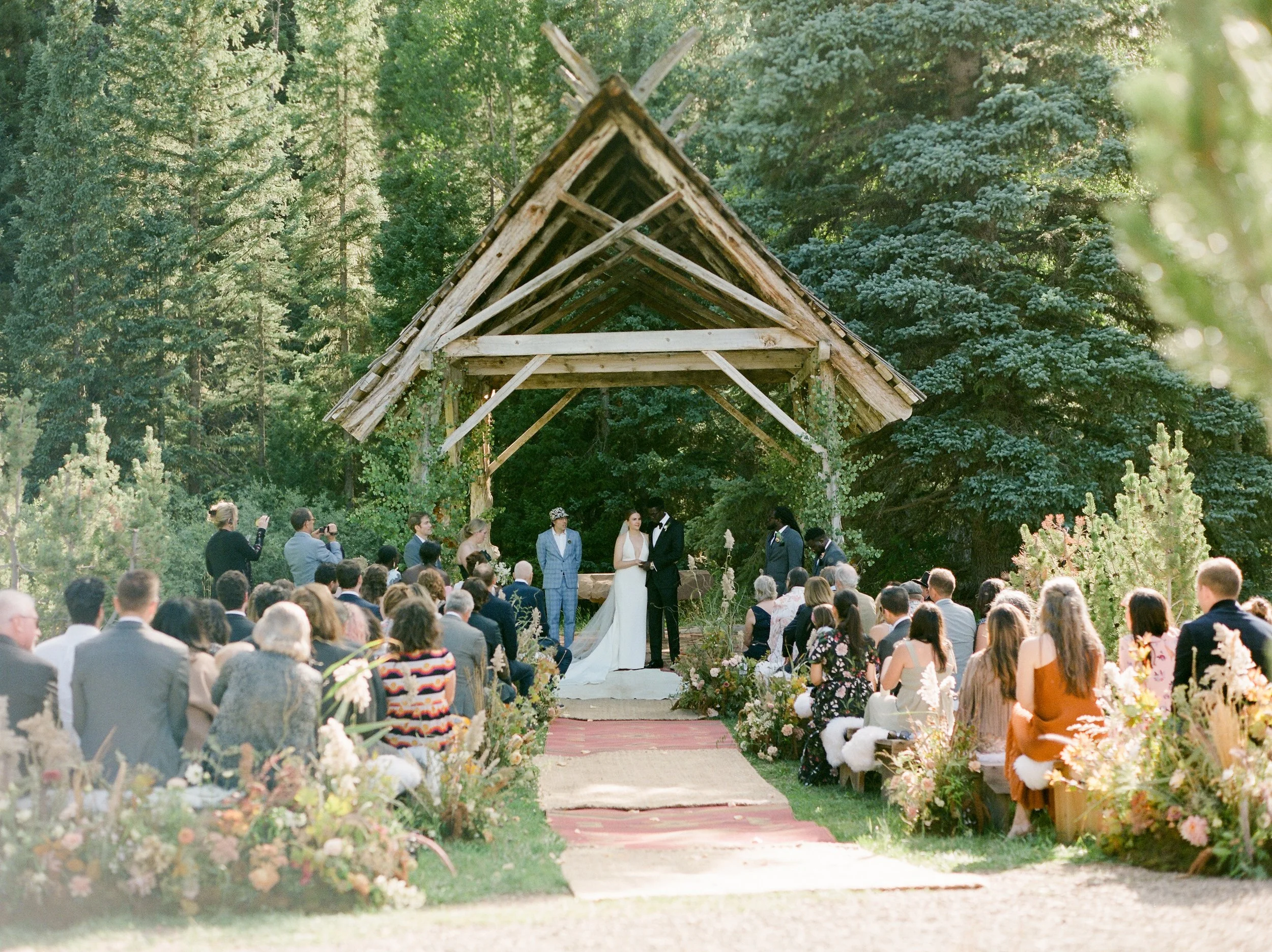 Outdoor wedding ceremony with a rustic wooden arch in a forest setting, bride and groom exchanging vows surrounded by seated guests and nature.