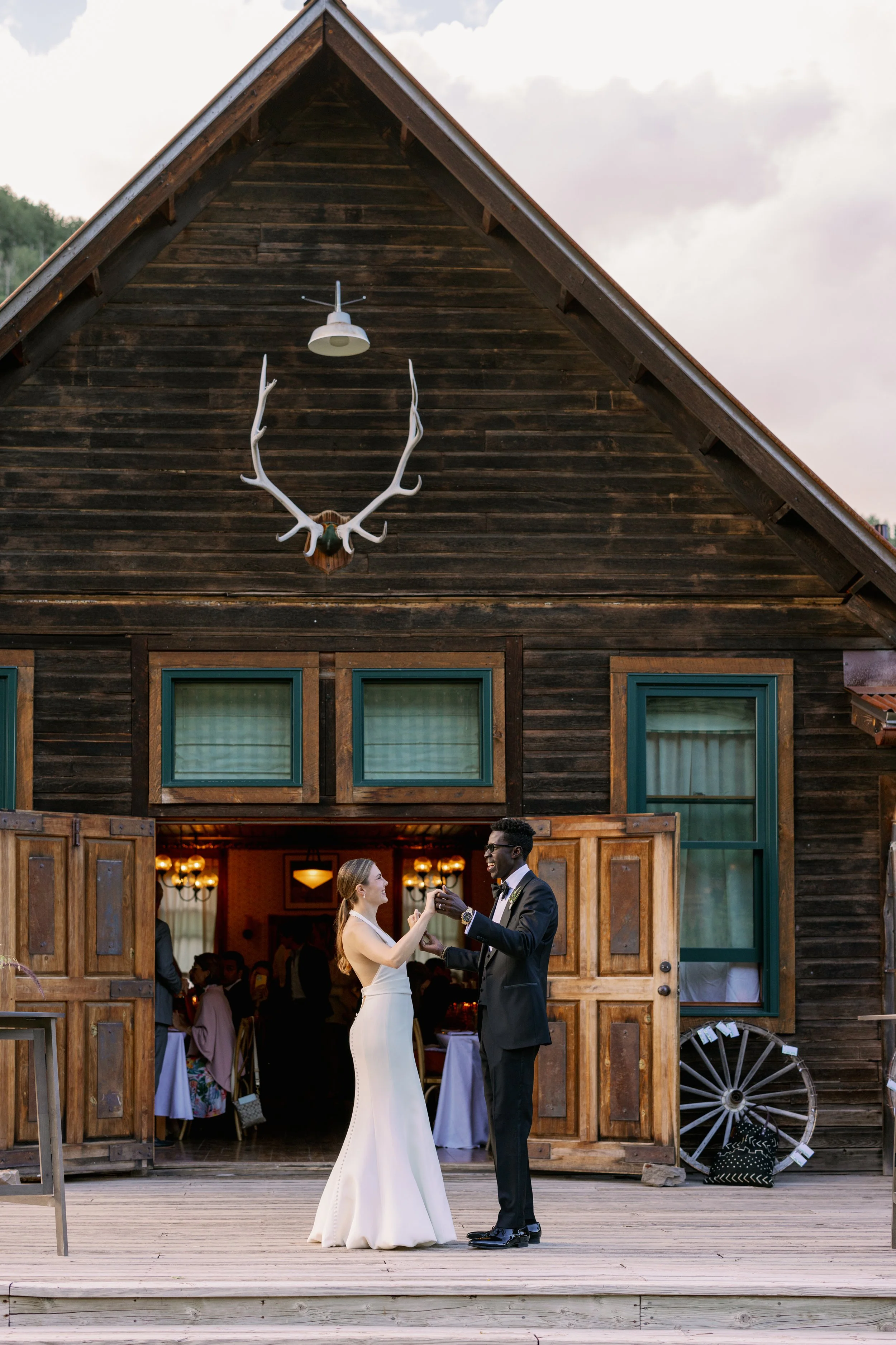 A bride and groom dancing outside a rustic wooden building with open doors, decorated with a mounted deer head and antlers.

The bride wears a white gown, and the groom is in a dark suit. They are smiling and holding hands, enjoying a moment together
