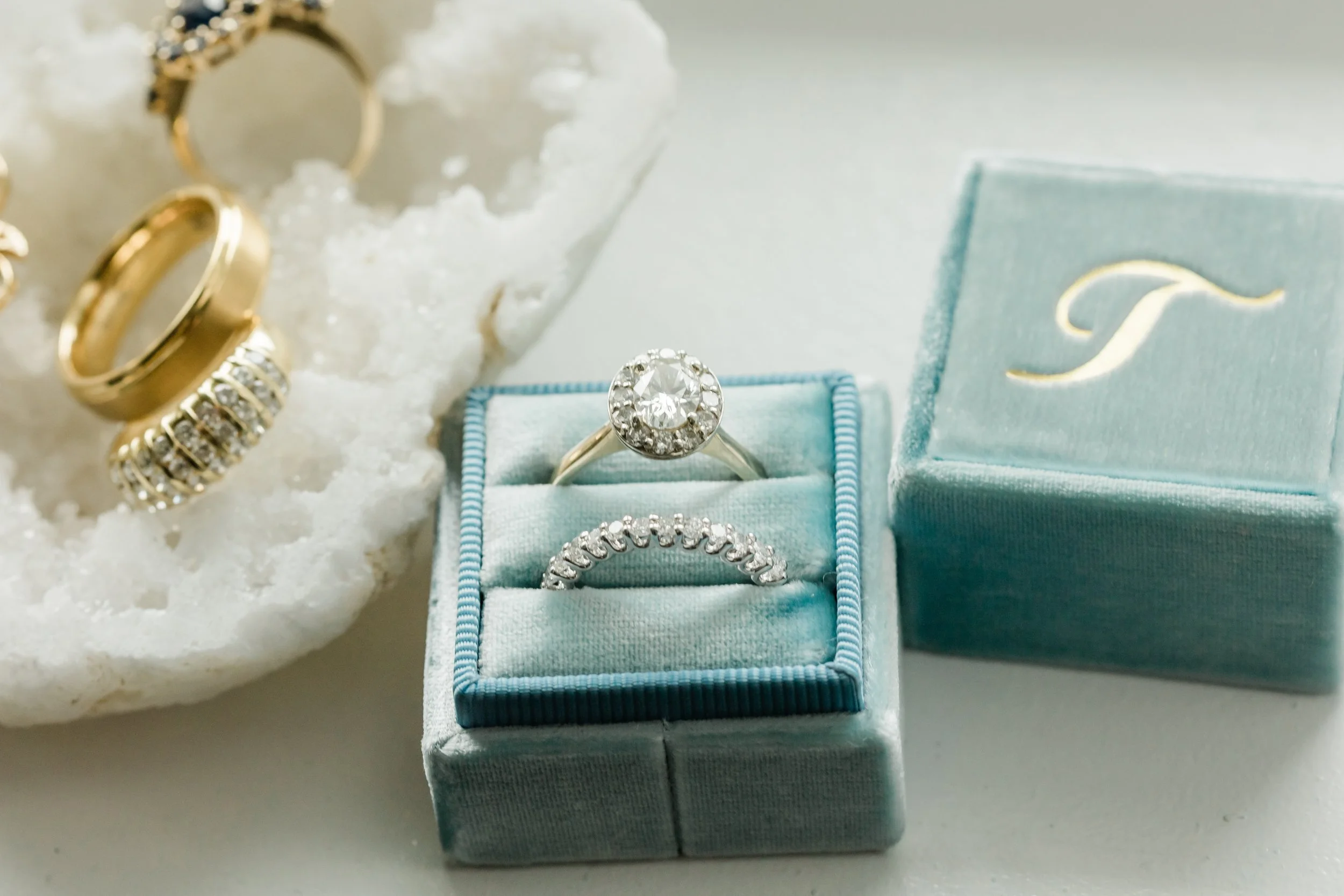 Diamond engagement ring and wedding band in a blue velvet jewelry box, with gold rings and jewelry on a white textured surface.