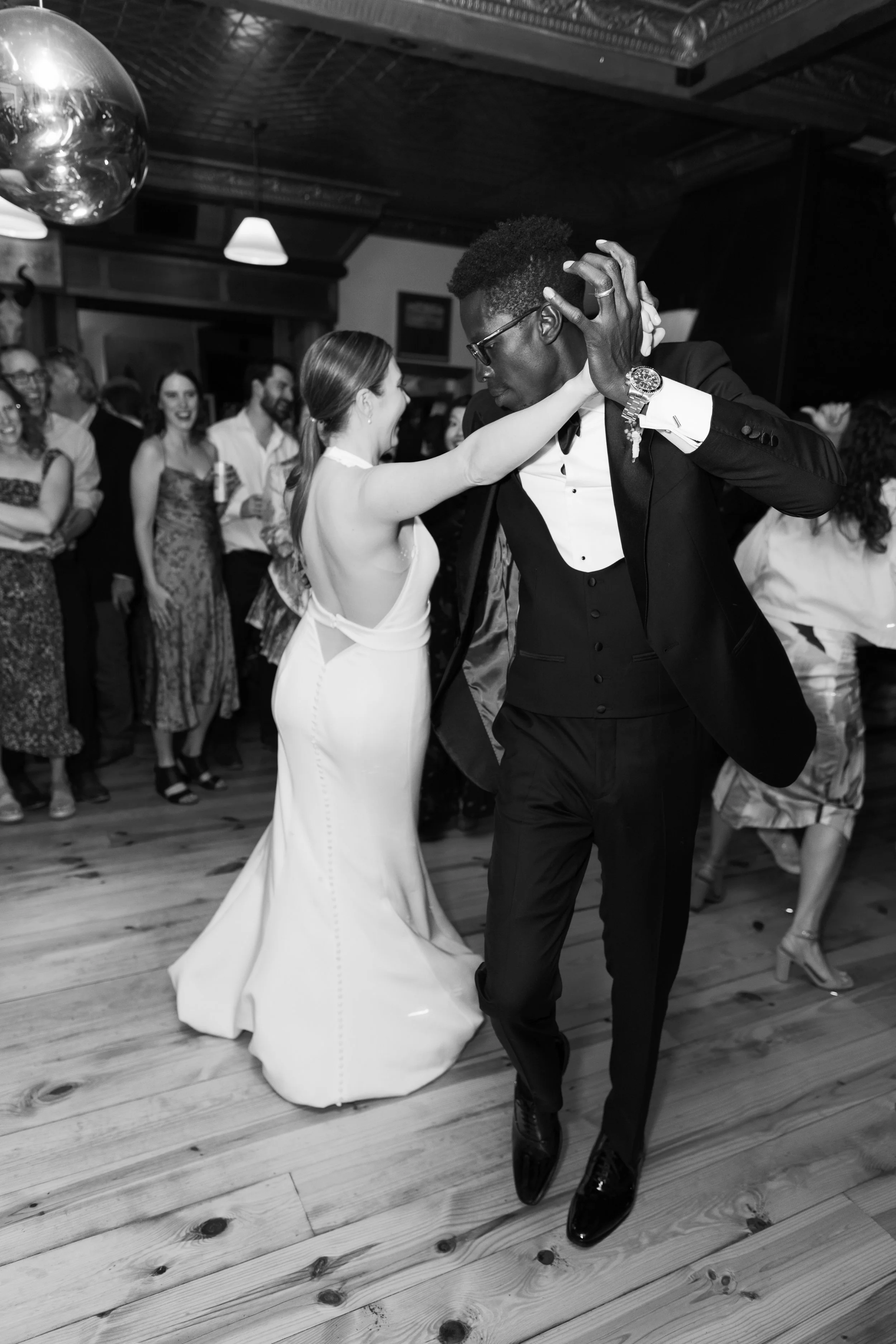 A black-and-white photo of a wedding reception with a bride and groom dancing. The bride is in a white wedding dress and the groom is in a black tuxedo. They are surrounded by guests enjoying the dance floor.