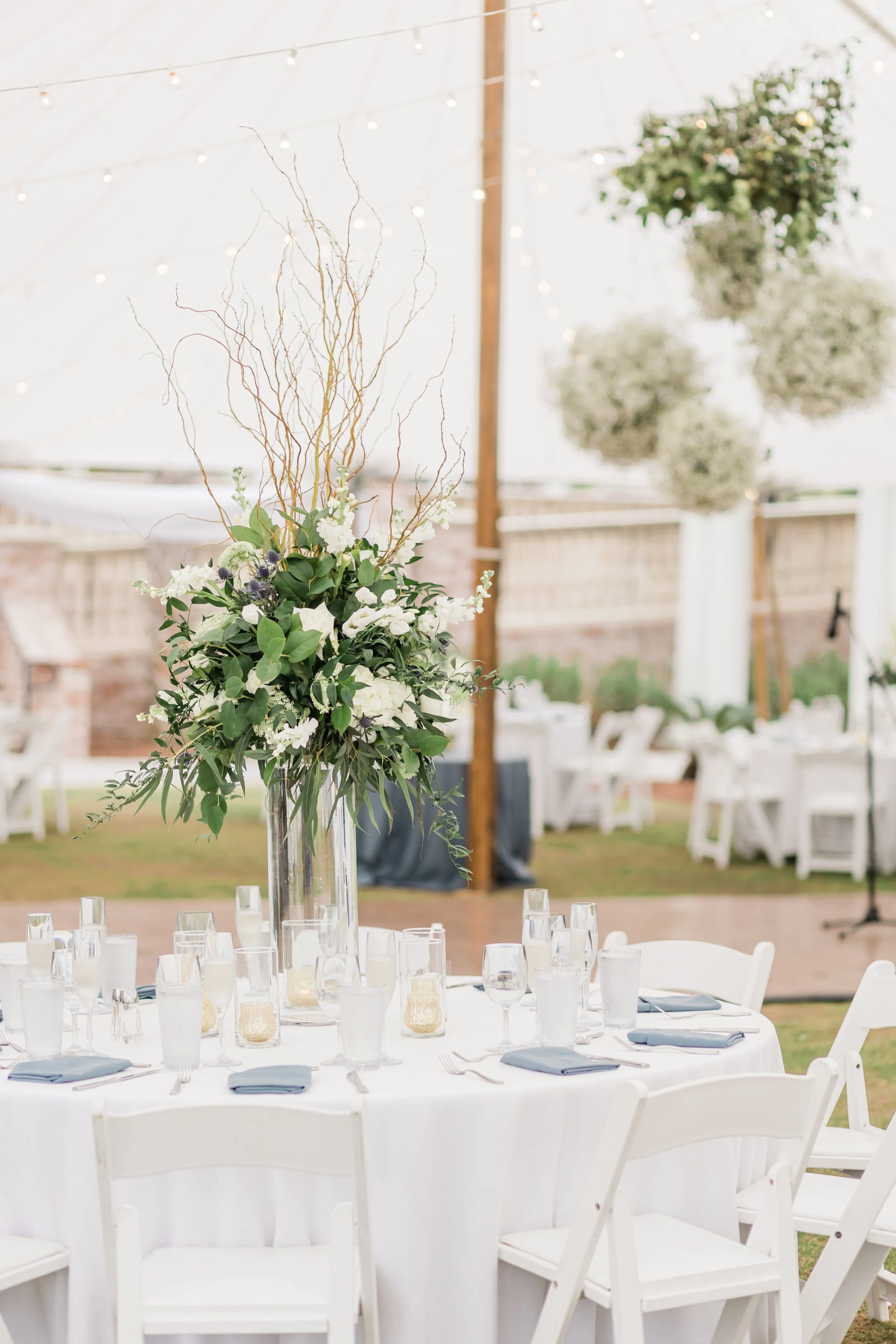 Wedding reception table with white tablecloth, tall floral centerpiece, and chairs outdoors decorated with hanging floral arrangements and string lights.