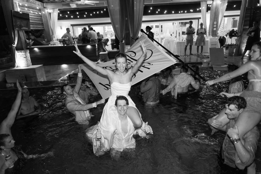 People celebrating in a swimming pool at a party, with some holding a sign and others cheering around.