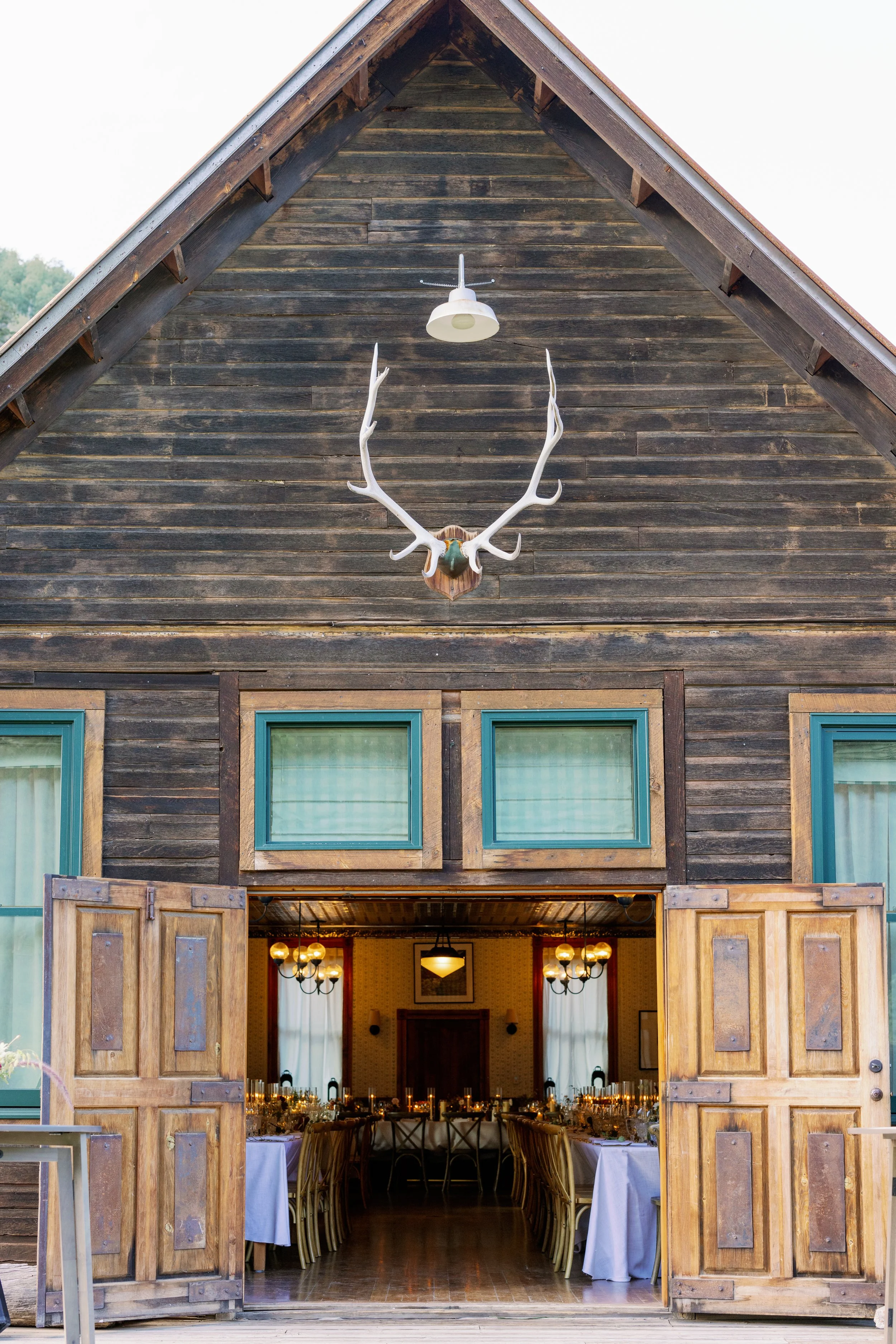 Open barn doors reveal a decorated dining hall with long tables, candles, and chairs inside a rustic wooden building, with antlers mounted on the exterior wall above.