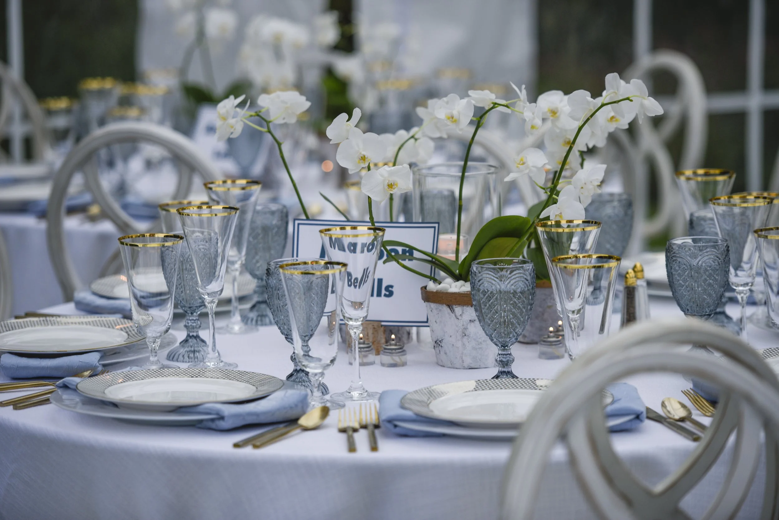 A formal outdoor dining table decorated with white orchids, glassware with gold rims, and gold utensils, set for an event or wedding reception.