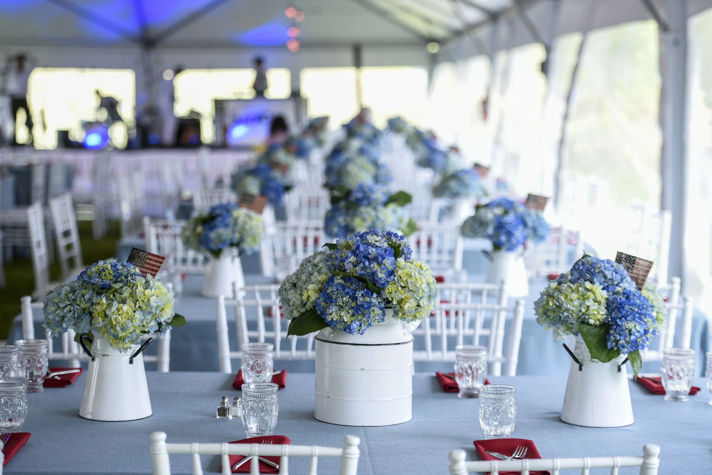 A banquet table decorated with blue and white flowers in white pitchers and a white box centerpiece in a tent with chairs, glasses, and red napkins.