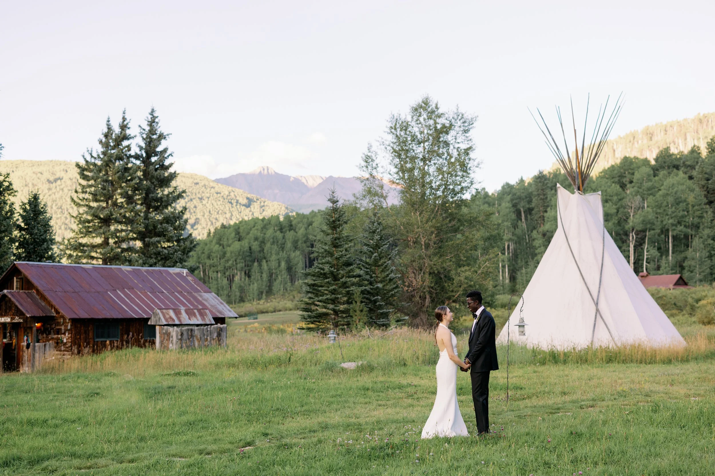 A bride and groom holding hands outdoors in front of a tipi tent, with a rustic barn, trees, and mountains in the background.