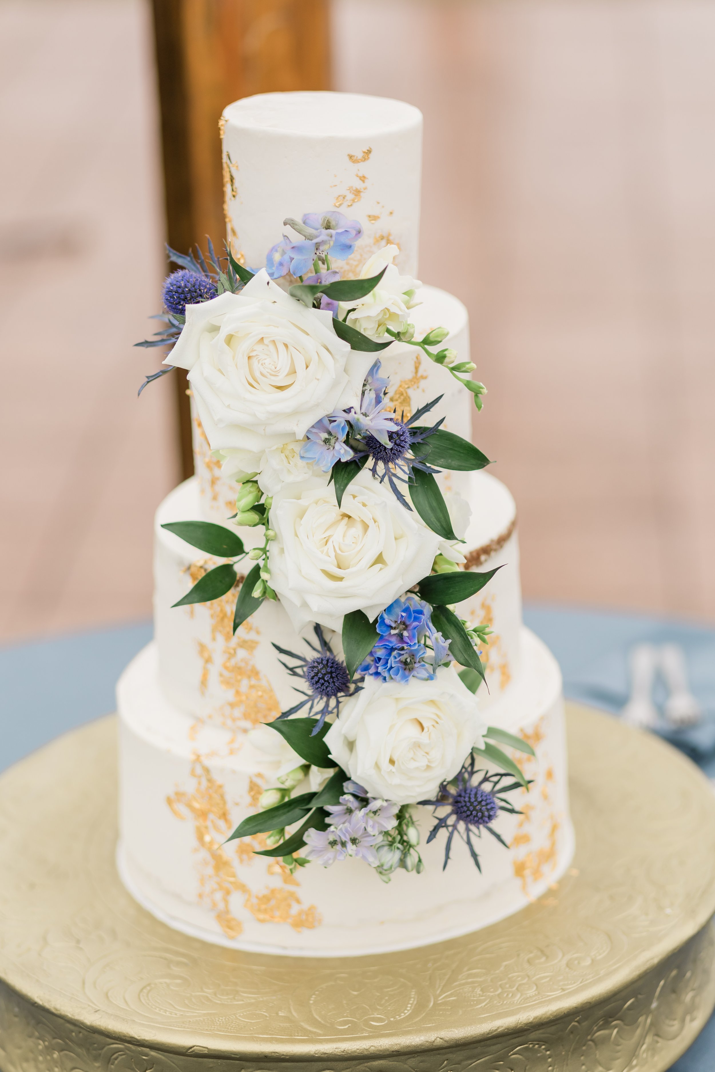 A multi-tiered wedding cake decorated with white roses, purple and blue flowers, and gold accents on a gold-colored stand.