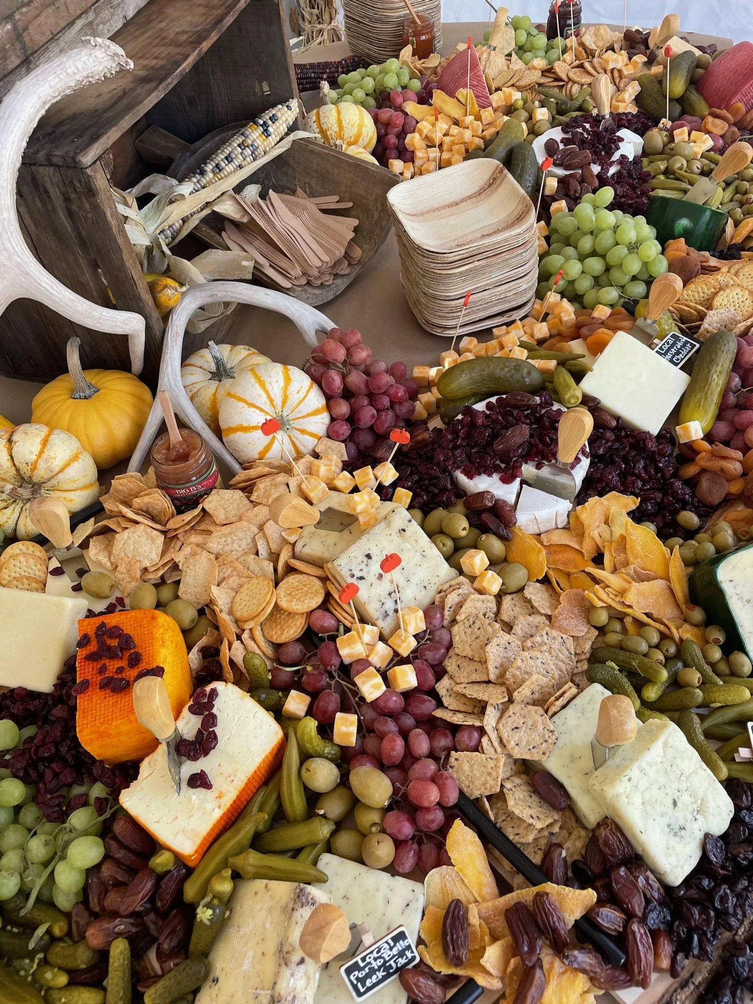 A display of assorted cheeses, grapes, crackers, and vegetables on a table, with some decorative pumpkin gourds and antlers.