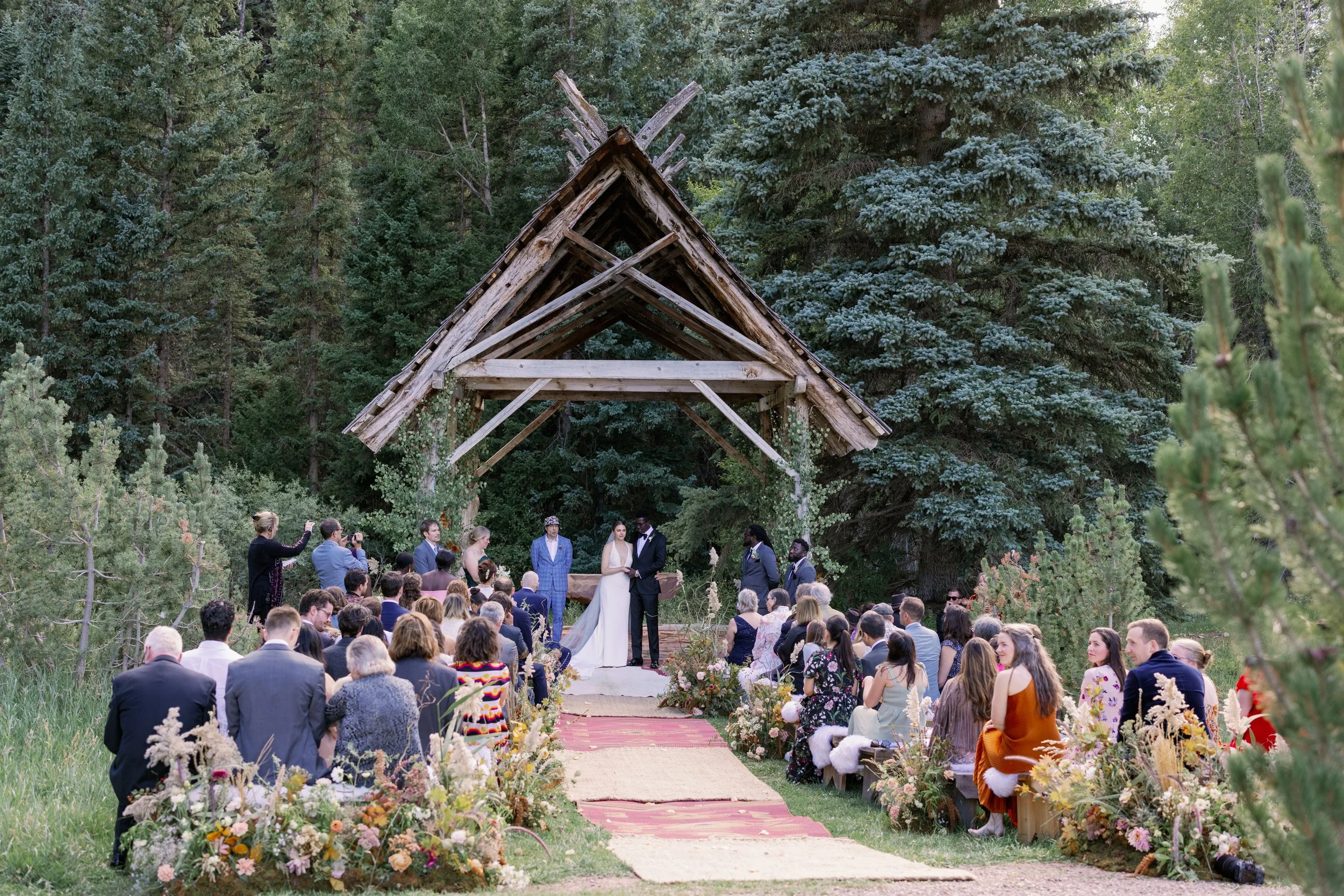 Outdoor wedding ceremony with a large gathering of guests seated on either side of a red carpet aisle, leading to a wooden arch where a bride and groom are exchanging vows, surrounded by lush green trees and floral arrangements.