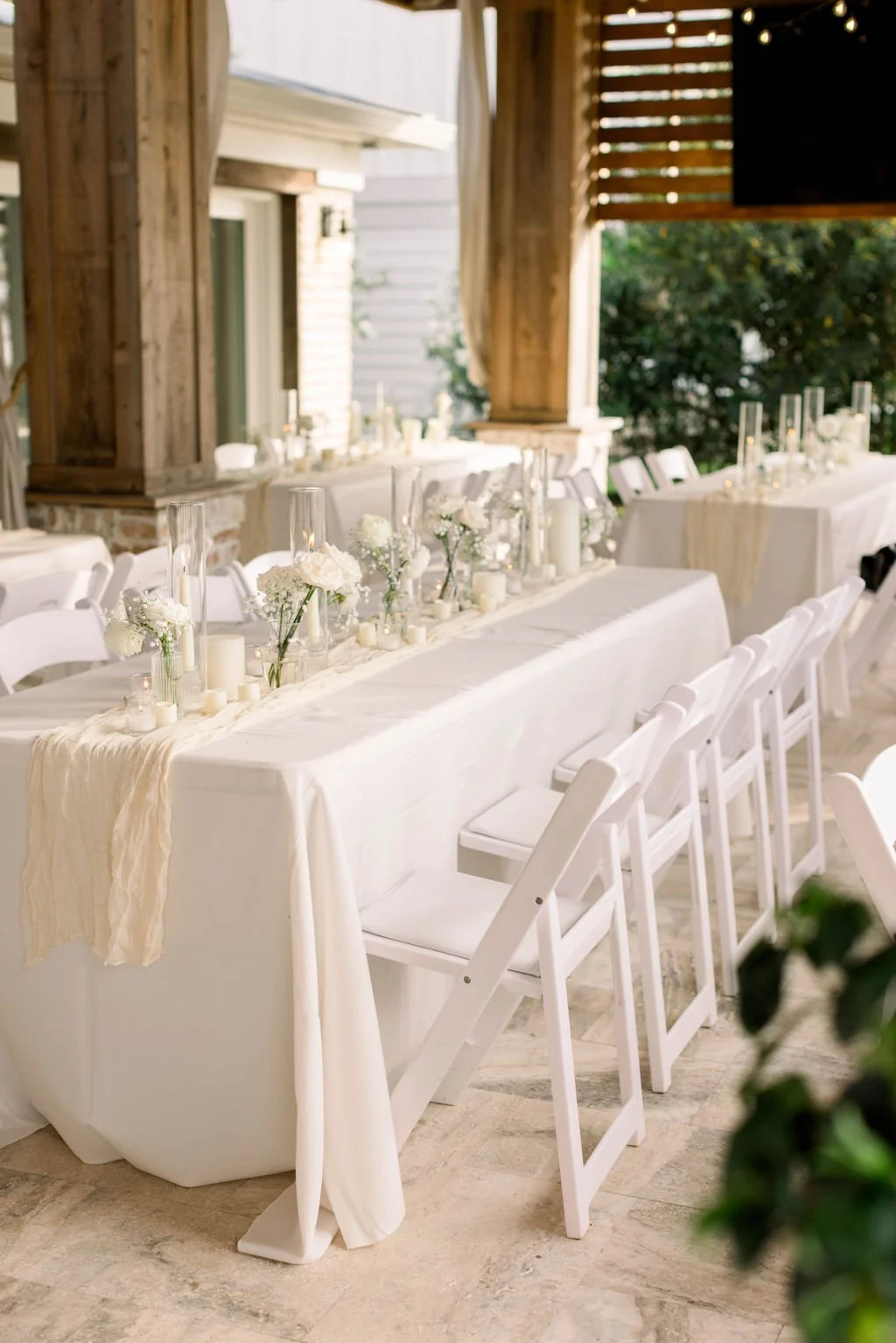 Elegant outdoor wedding reception table decorated with white flowers, candles, and tall glass vases, set in a rustic pavilion with wooden beams and natural light.