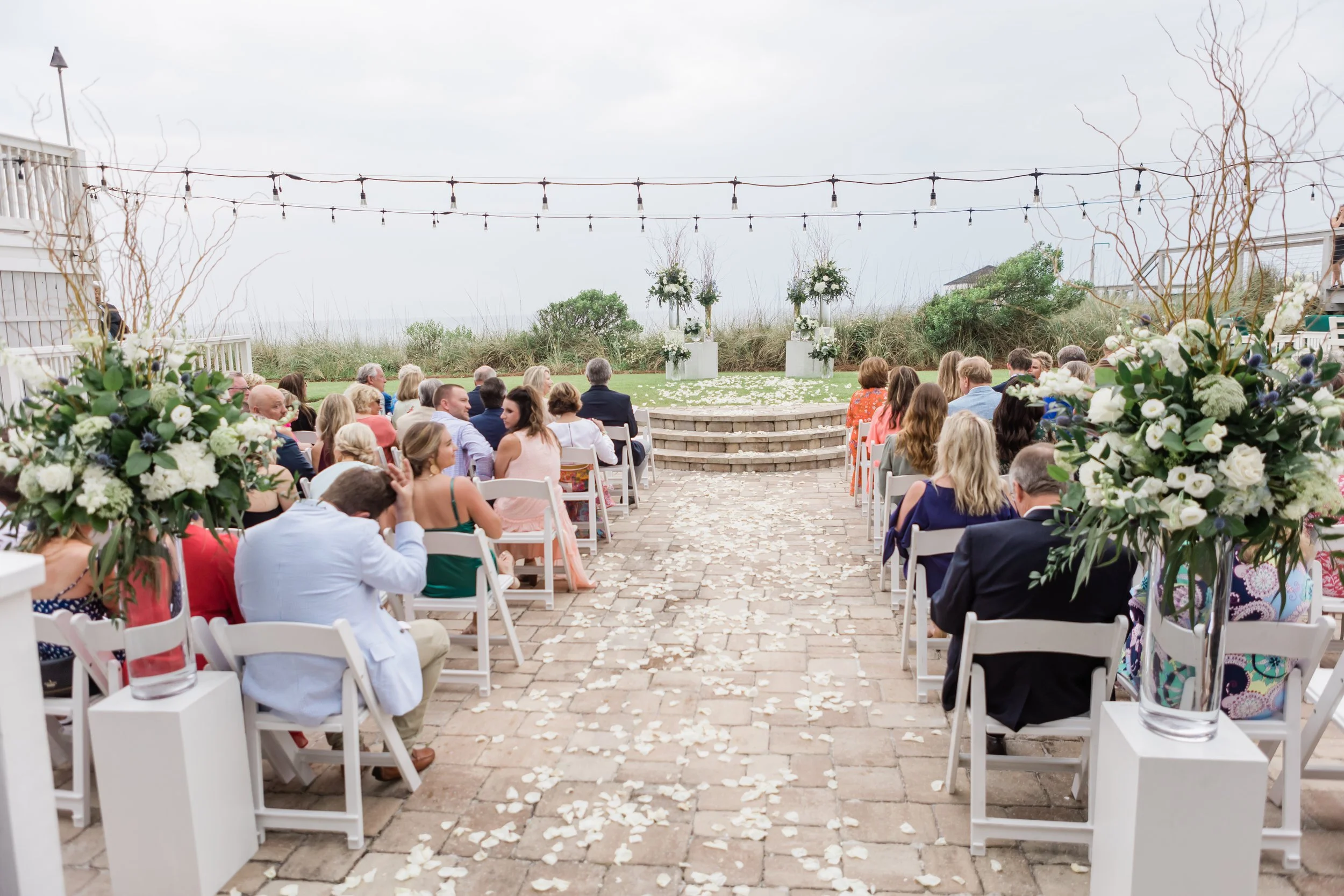Outdoor wedding ceremony with chairs, flower arrangements, and rose petal aisle, set on a stone patio overlooking a natural landscape.
