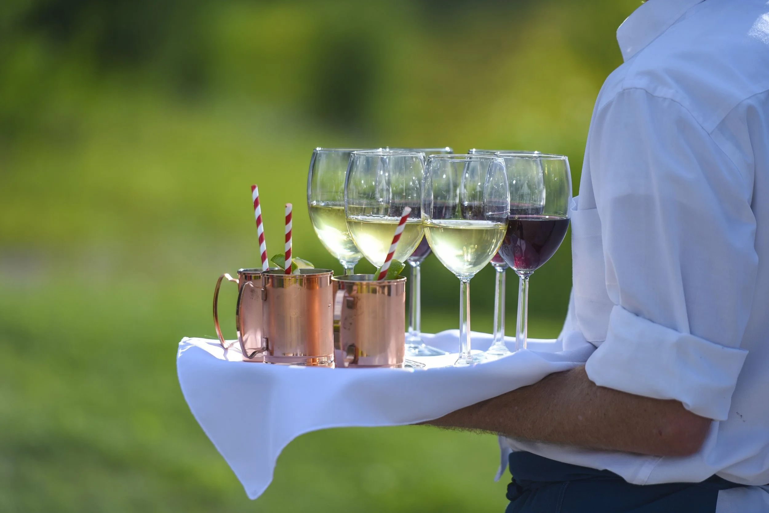 A person holding a white tray with several glasses of wine and copper cups with striped paper straws, set outdoors on a grassy area.