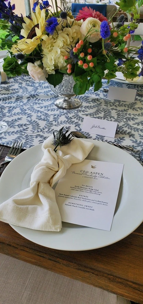 Elegant table setting with a white plate, cloth napkin tied with a feather, menu card, and a floral centerpiece with white, pink, purple, and blue flowers in a glass vase.