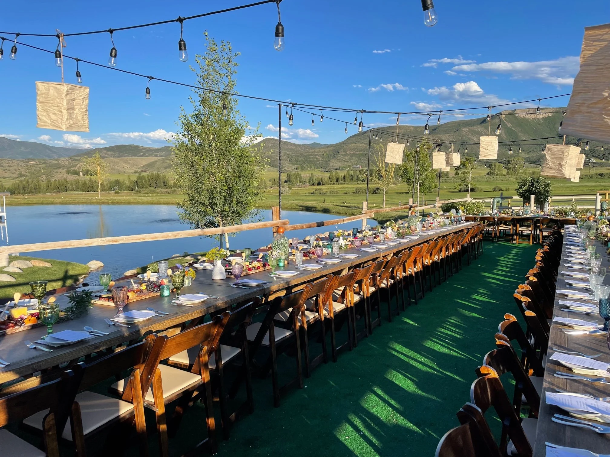 Outdoor dining setup on a long wooden table with chairs, decorated with flowers and tableware, set beside a lake with mountains in the background, under string lights on a sunny day.