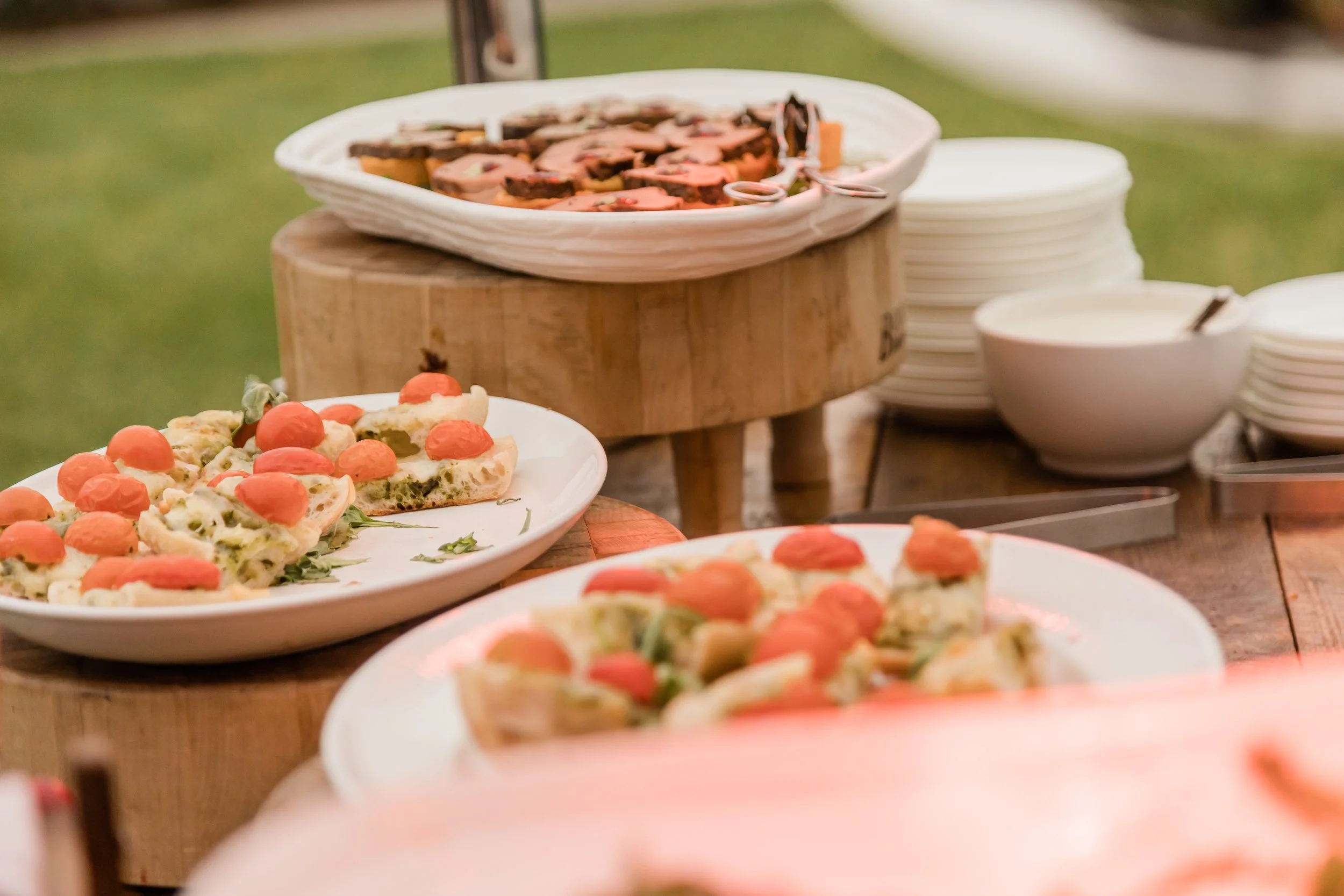Plates of sliced Italian pizza with cherry tomato toppings on a wooden table, with bowls of sliced pizza in the background.