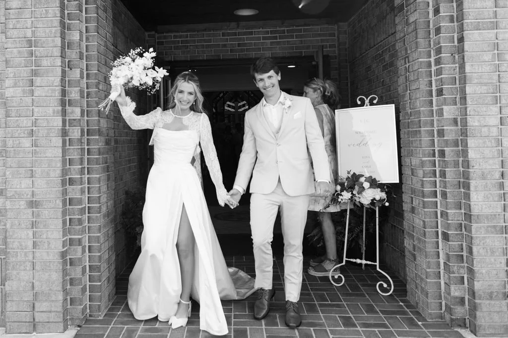 Bride and groom leaving a wedding reception, holding hands, with the bride waving and holding a bouquet. The bride is in a long white wedding dress with lace sleeves, and the groom is in a white suit. They are smiling and walking through a brick arch