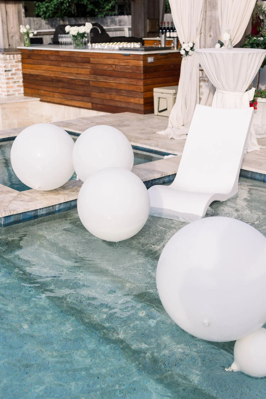 A poolside scene with a white lounge chair, large white globe-shaped decorations in the water, and a wooden bar area with white curtains and flowers in the background.